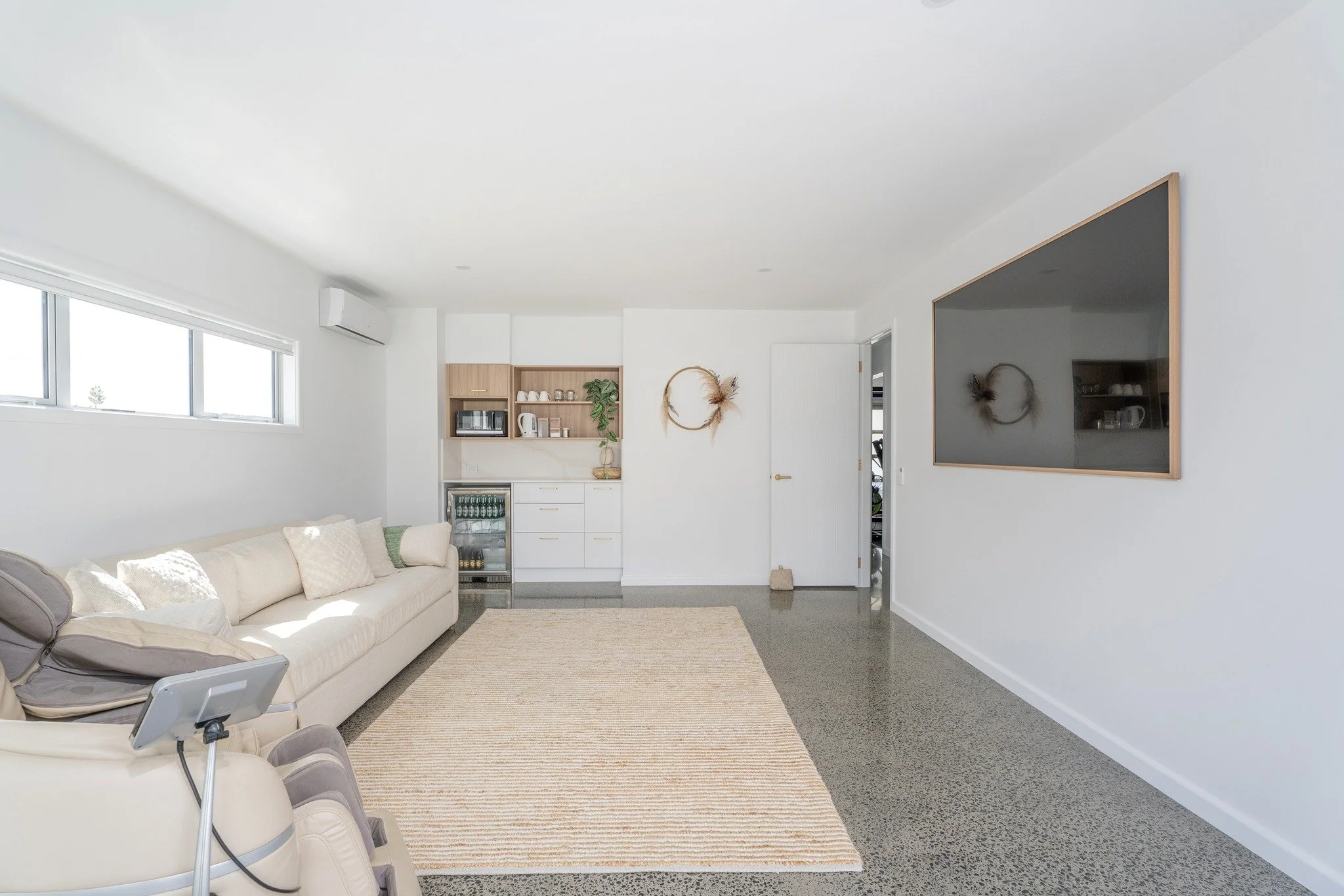 Minimalist living room with beige sofa and recliner, window with blinds, mounted TV, small kitchenette with open shelves, countertop with a few items, and wall decor with circular wreaths, on polished concrete floor with a beige rug.