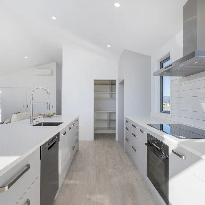 Modern white kitchen with built-in appliances, a sink, and open shelving, featuring windows and recessed lighting.
