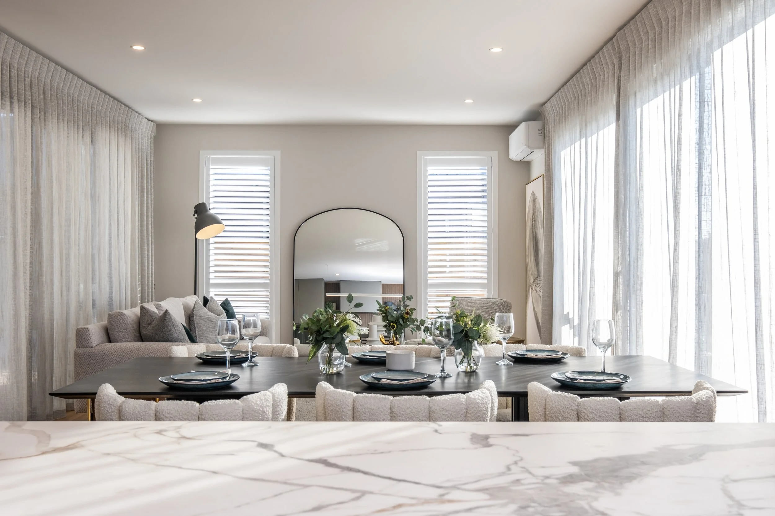 Contemporary dining room with a large black table set with wine glasses and plates, surrounded by cream-colored chairs, and decorated with green plant centerpieces, with white curtains and a mirror on the wall.