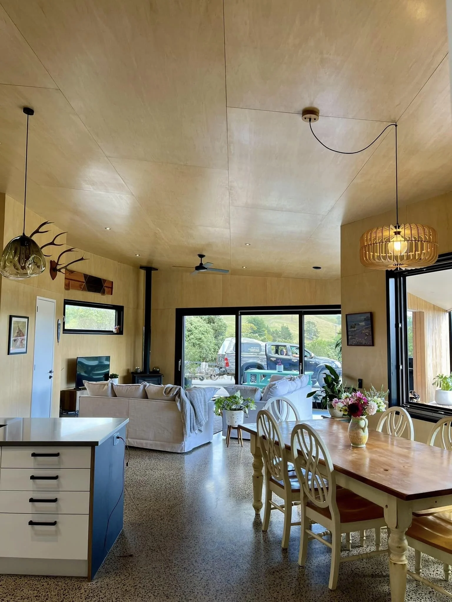A modern open-concept living and dining area with wood-paneled walls and ceiling, large windows, a white sofa with a blanket, potted plants, a wooden dining table with chairs, and decorative lighting fixtures.