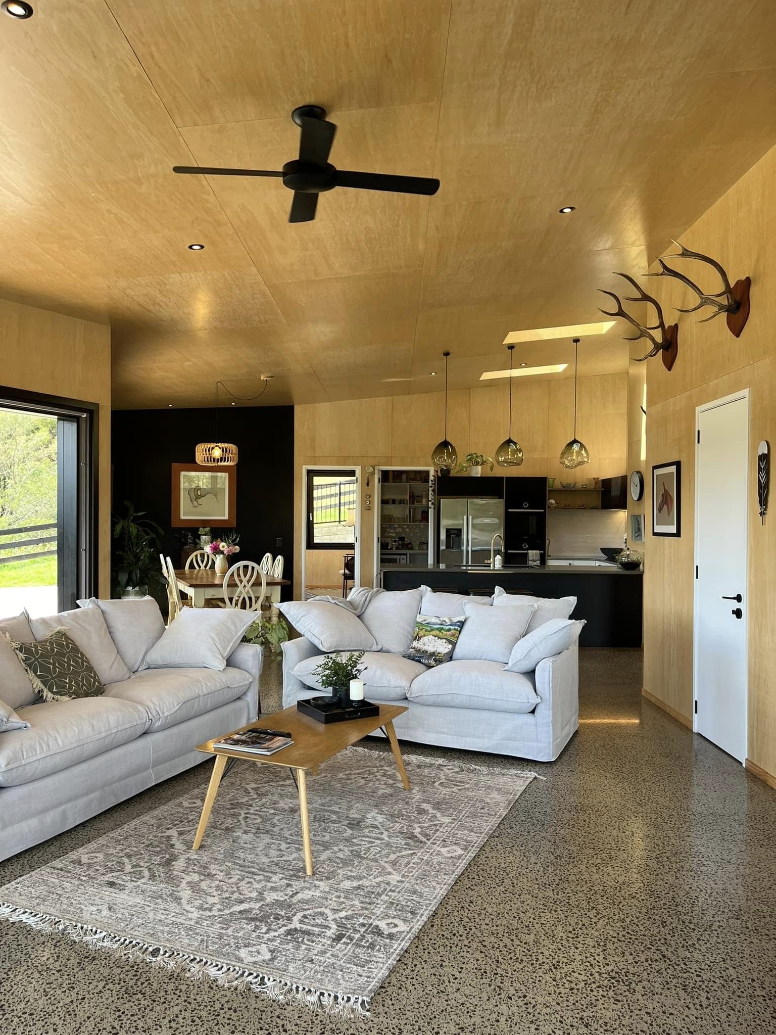 Open-plan living room and dining area with white sofas, wooden coffee table, and black ceiling fan, featuring wooden walls, black kitchen cabinets, and decorative mounted antlers.