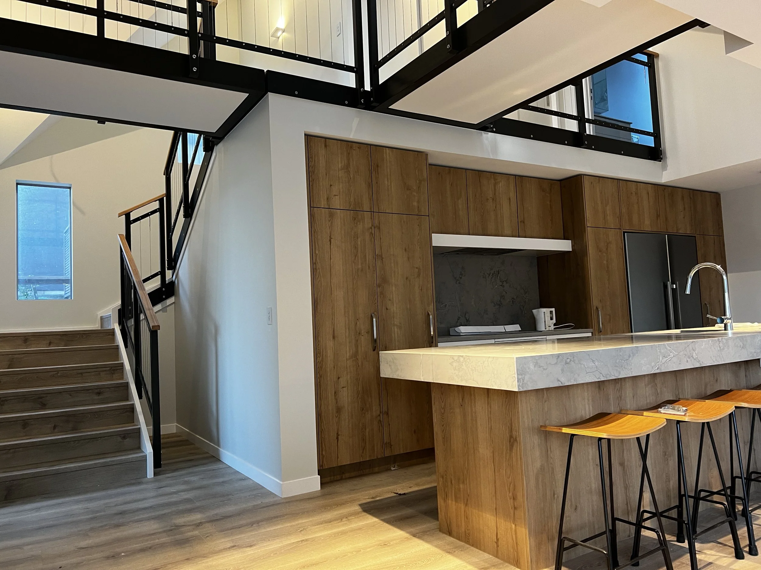 Modern kitchen with a beige marble island, wooden cabinetry, a stainless steel refrigerator, and a loft area with black metal railings, wooden staircase, and hardwood floors.