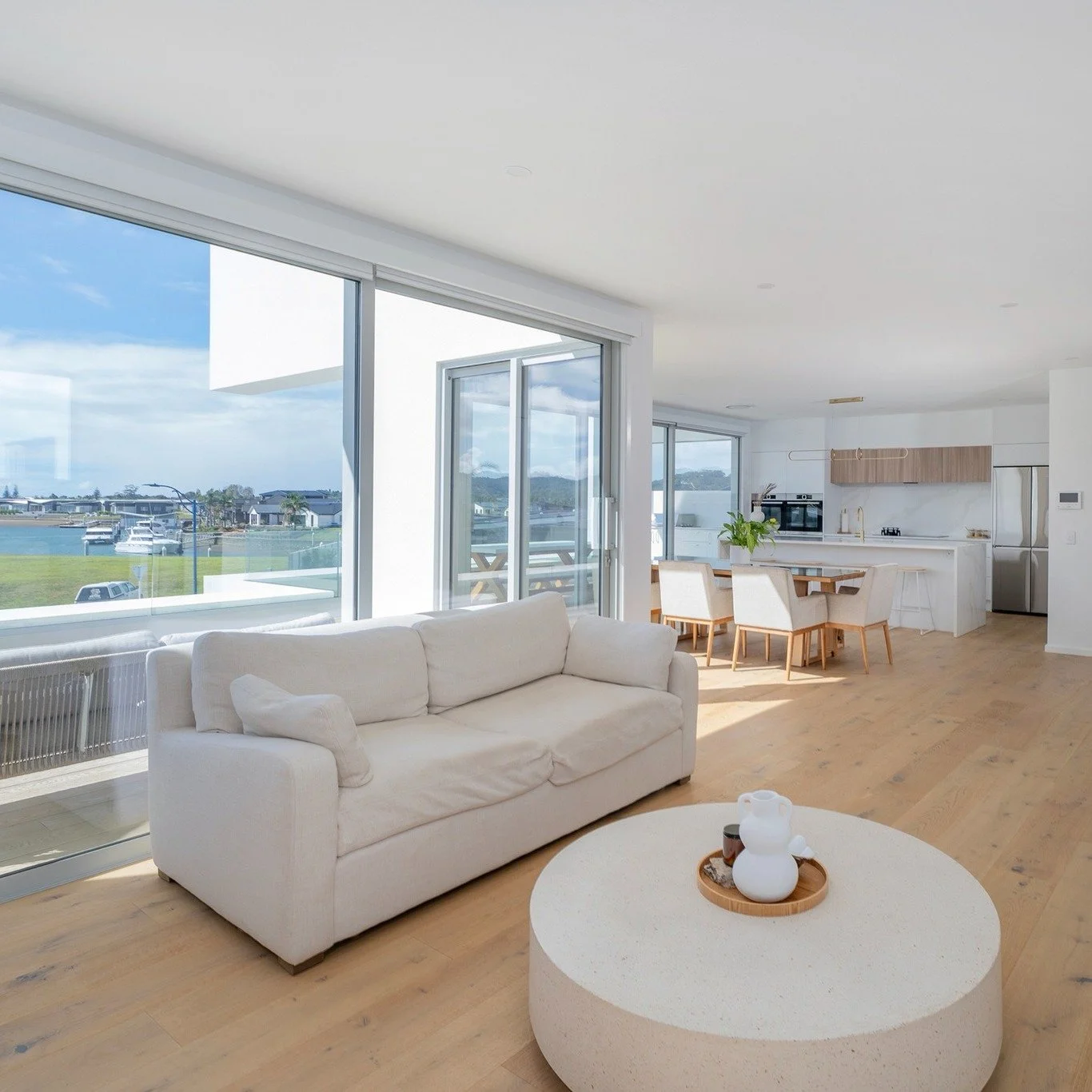 Bright, modern open-concept living room and kitchen with large windows, white sofa, round coffee table, and wooden dining table with six chairs.