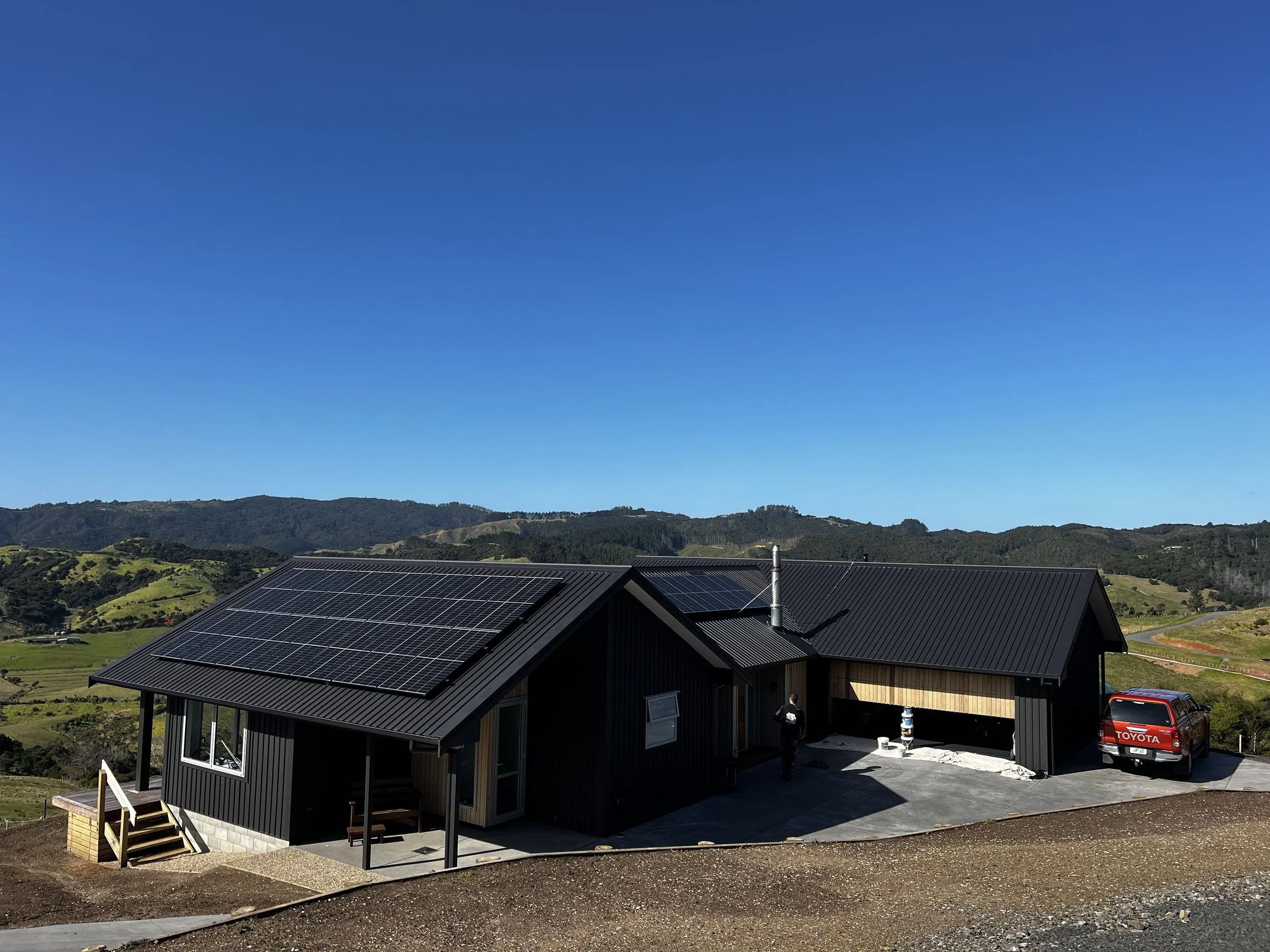 Modern house with solar panels on roof, black exterior, concrete driveway, and a red Toyota truck parked outside, set in a mountainous landscape with clear blue sky.