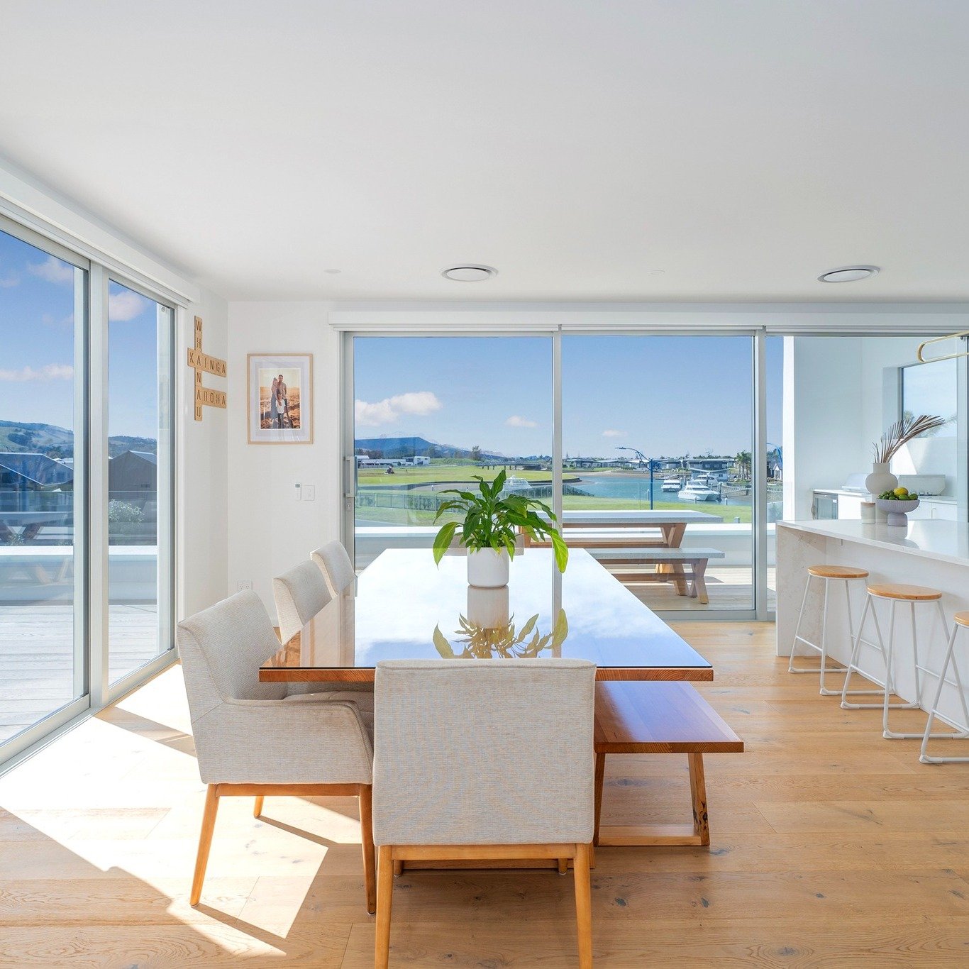 Bright dining area with large glass sliding doors and windows overlooking a marina with boats and green fields. There is a wooden dining table with six light-colored chairs, a potted plant in the center, and a balcony outside with a picnic table. The