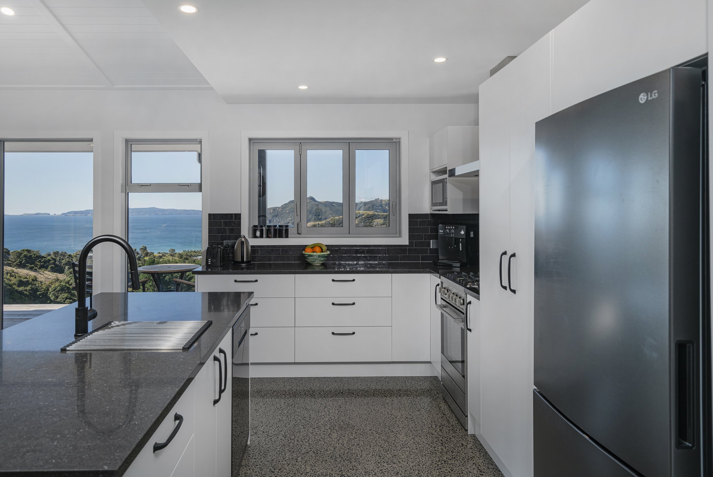 Modern kitchen with white cabinets, black countertops, stainless steel appliances, and a view of the ocean and distant mountains through the windows.