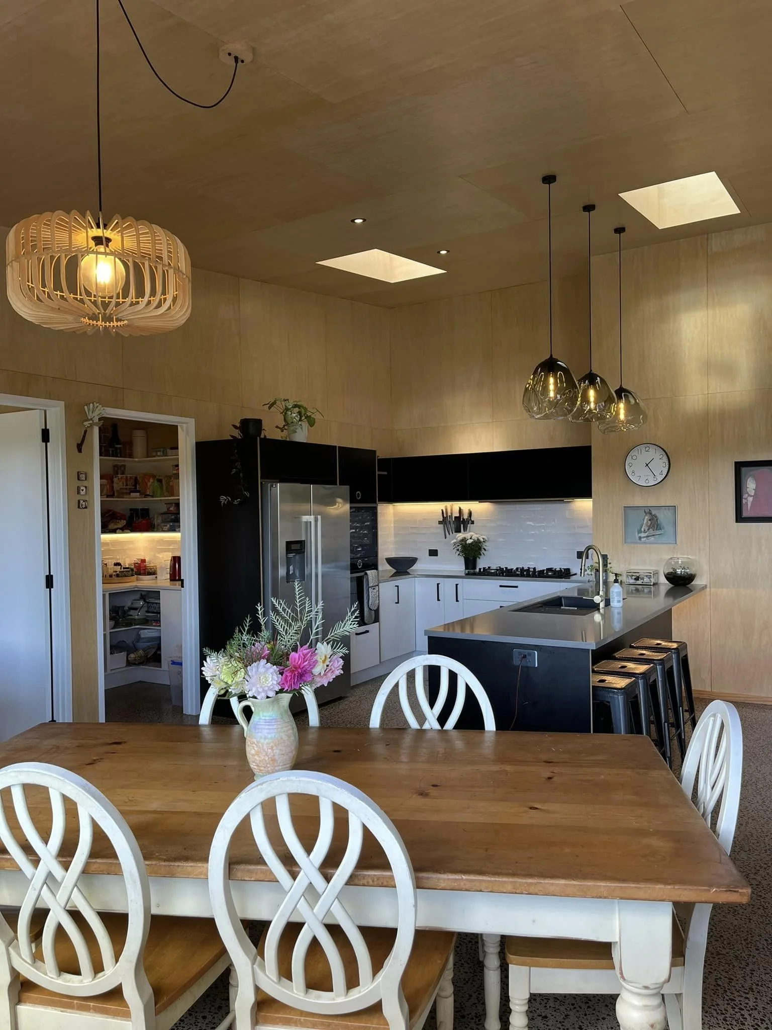 A modern kitchen and dining area with a wooden table and white chairs. The kitchen features black and white cabinets, a stainless steel refrigerator, a small sink, and hanging pendant lights. There is a vase with pink and white flowers on the table, 
