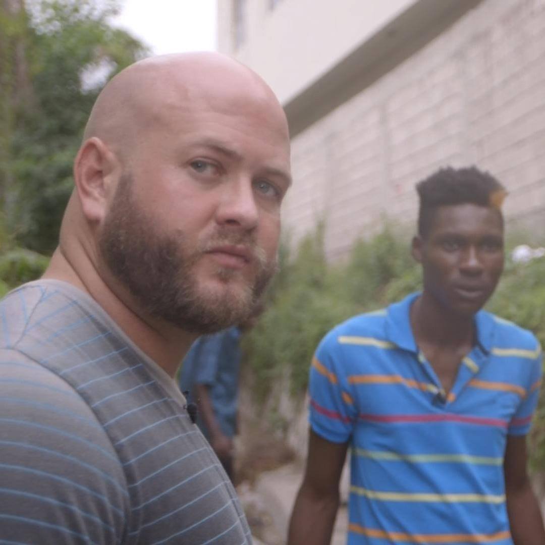 Mitchell Williams, a bald man with a beard in a striped shirt looking at the camera, standing outdoors with another young man of Jamaican and African descent in a striped shirt in the background.