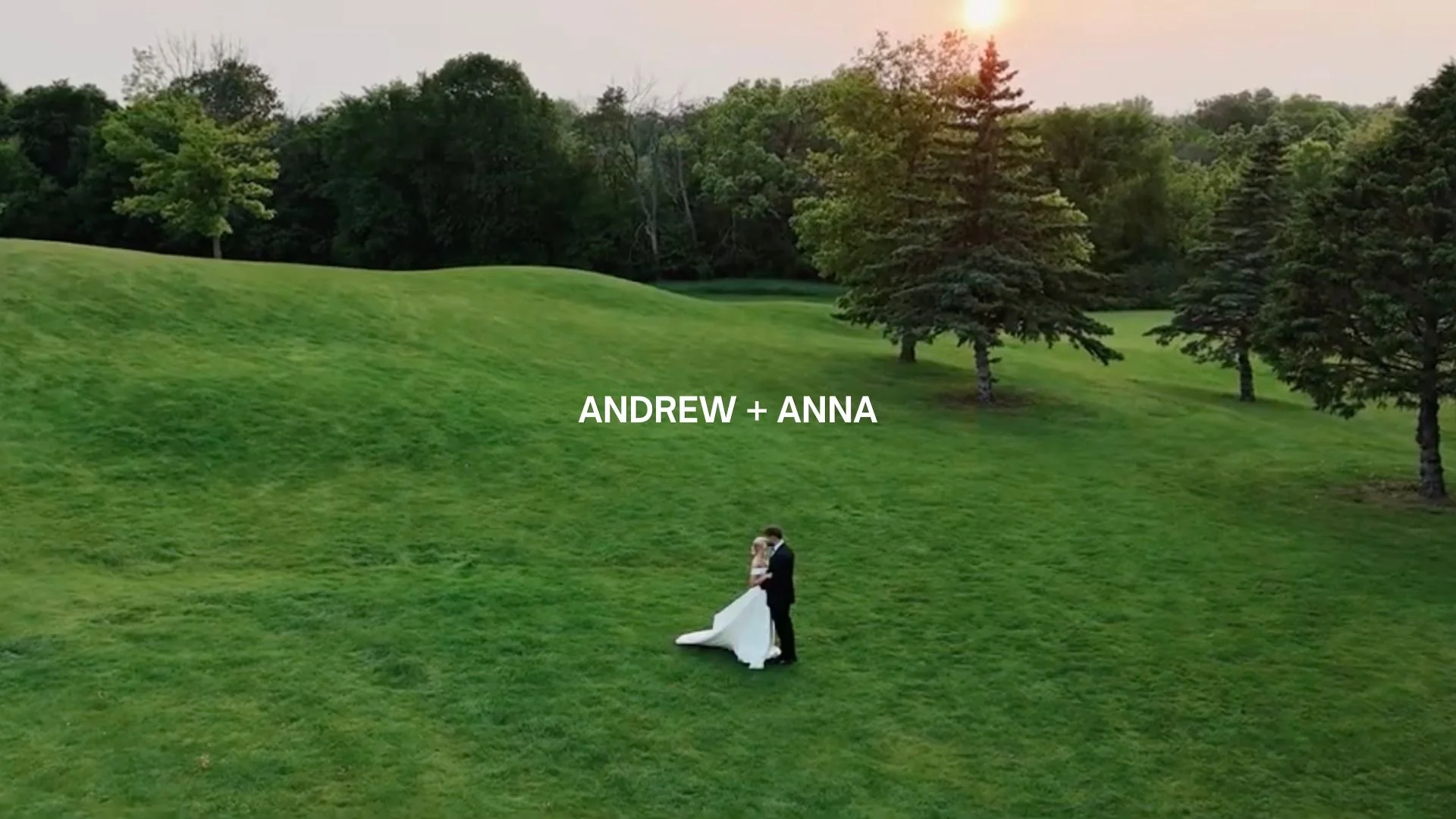 A bride and groom are standing on a grassy hilltop surrounded by trees at sunset, with the text 'Andrew + Anna' overlayed in the center.