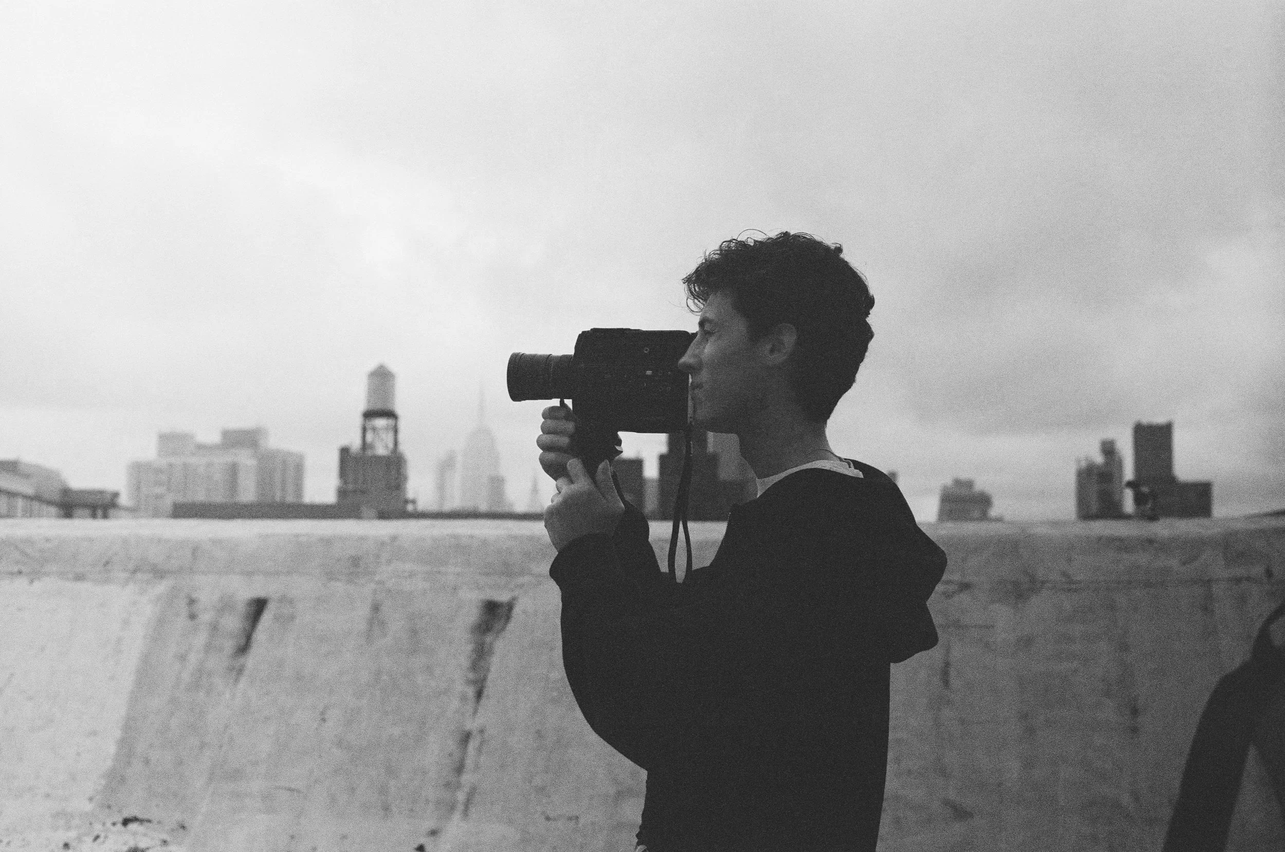 Black and white photo of a young man with curly hair using a video camera on an outdoor rooftop, with city buildings in the background under an overcast sky.