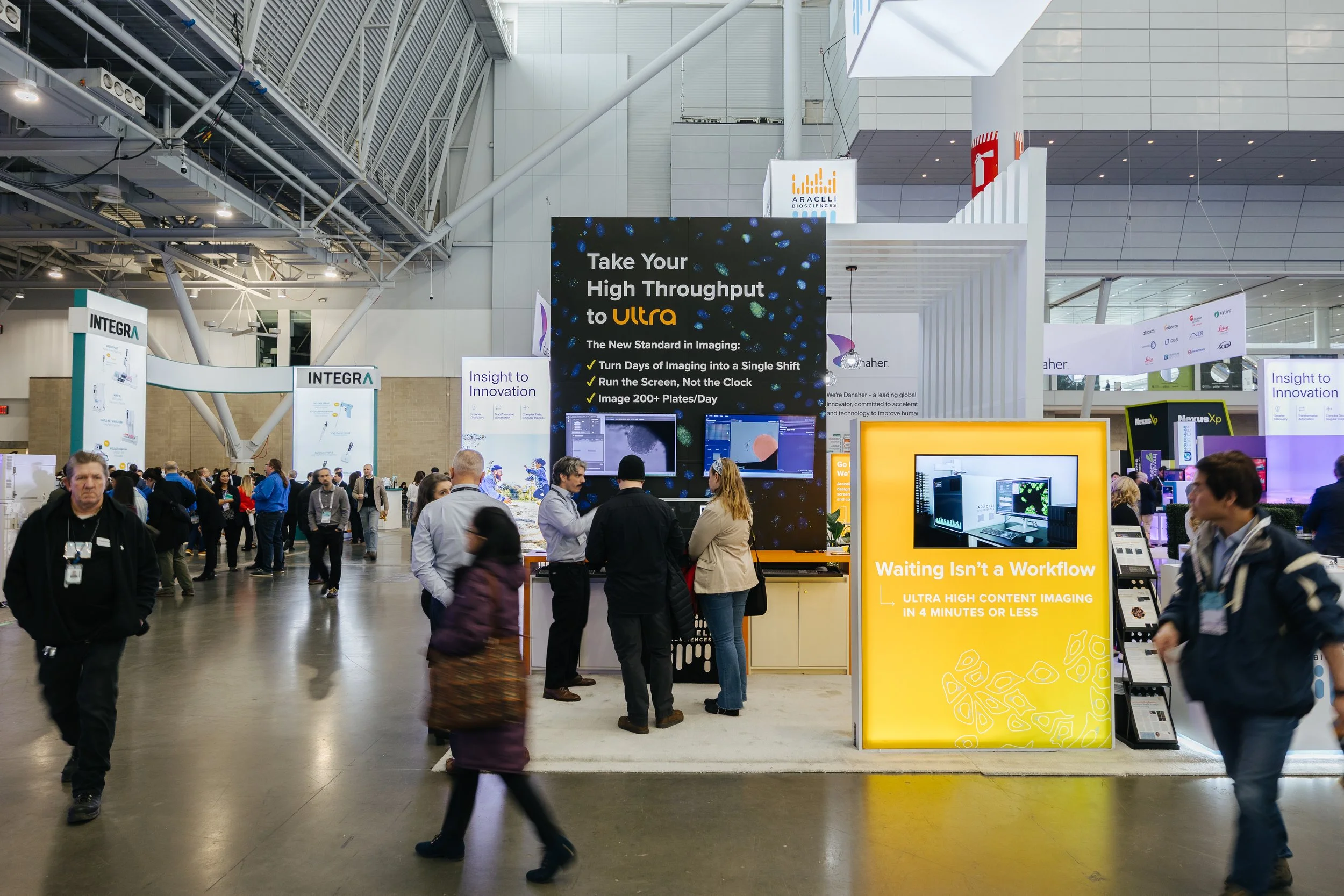 A busy trade show or conference with people walking and standing around exhibitor booths. Prominent signs include one advertising high throughput imaging technology and another with the slogan "Waiting Isn't a Workflow". The setting is an indoor convention center with high ceilings, metal and glass architecture, and various company logos visible.