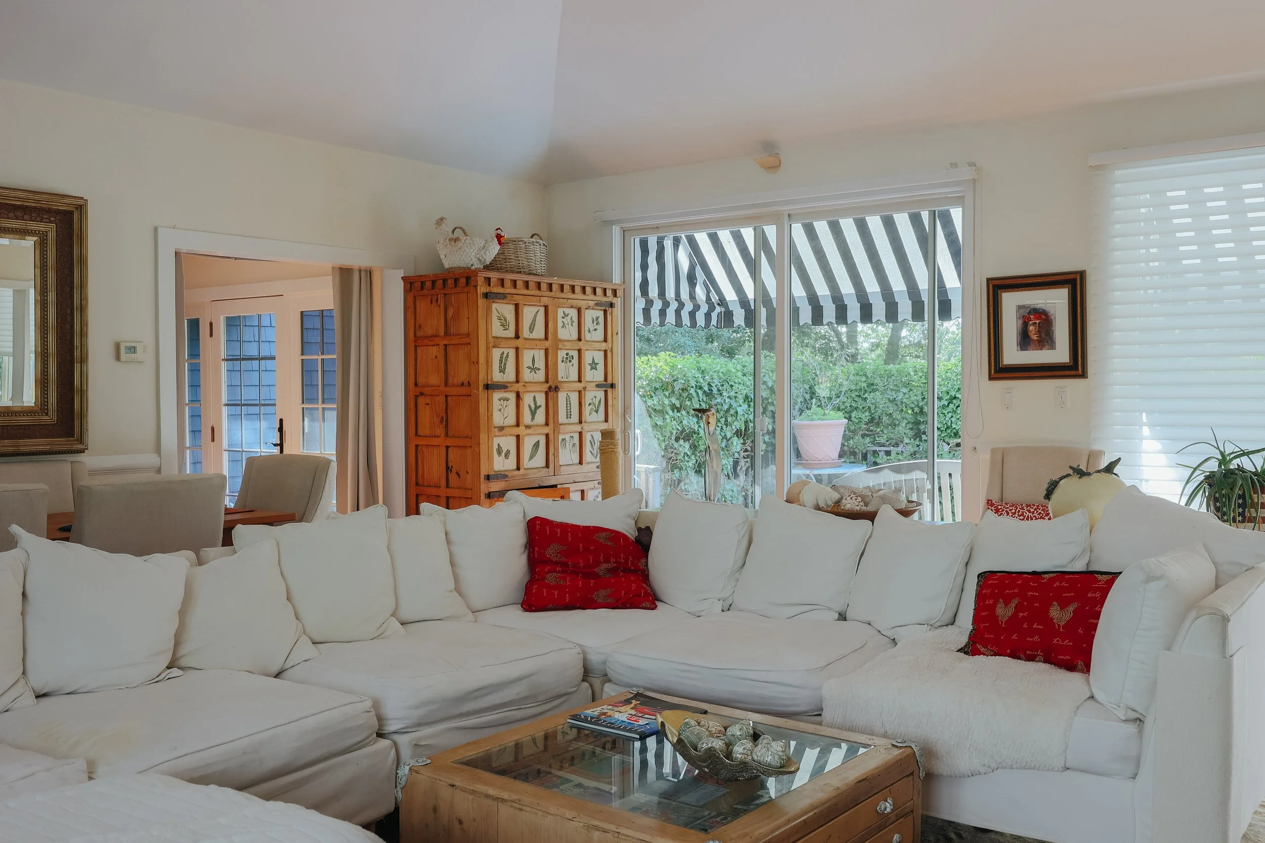 A living room with a large white sectional sofa and red pillows, a glass-topped wooden coffee table with seashells, a wooden cabinet with botanical artwork, and large sliding glass doors opening to a green outdoor area.