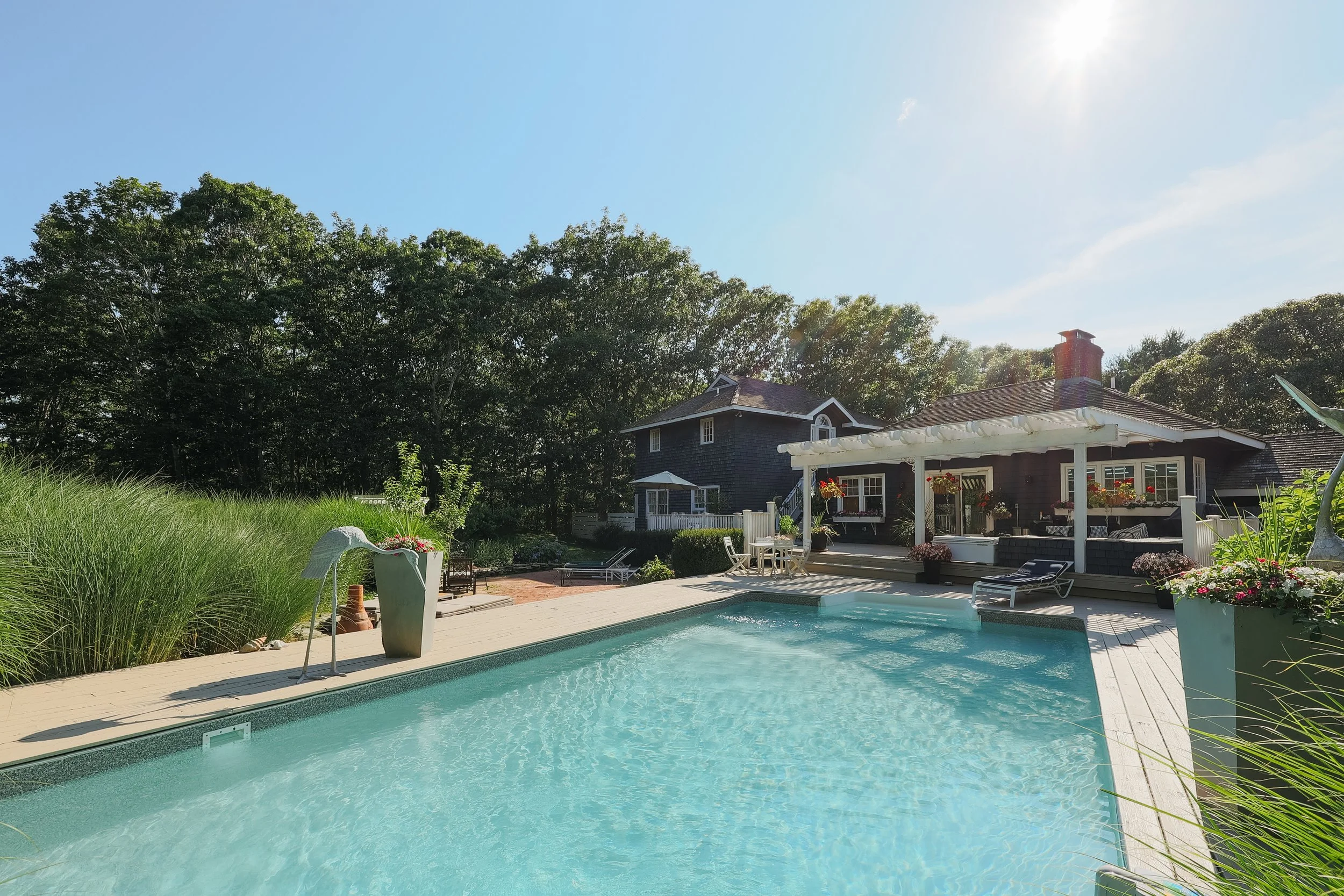 A backyard with a swimming pool, a wooden deck, and a house with a porch. There are outdoor furniture, potted plants, and lush greenery, with trees in the background under a clear blue sky.