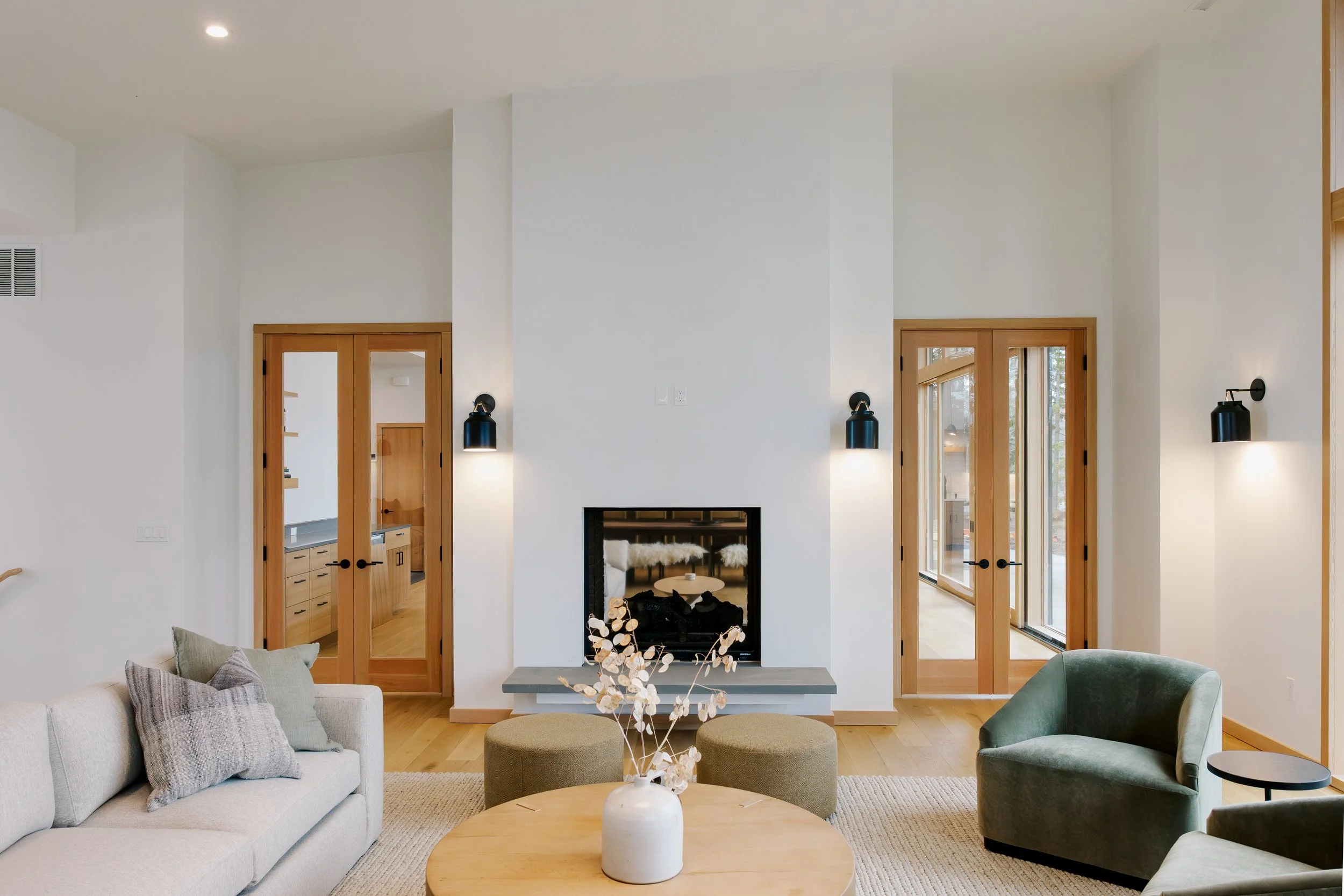 Living room with a white sofa, green armchair, round coffee table with a white vase, natural wood flooring, and large windows with wooden framing.