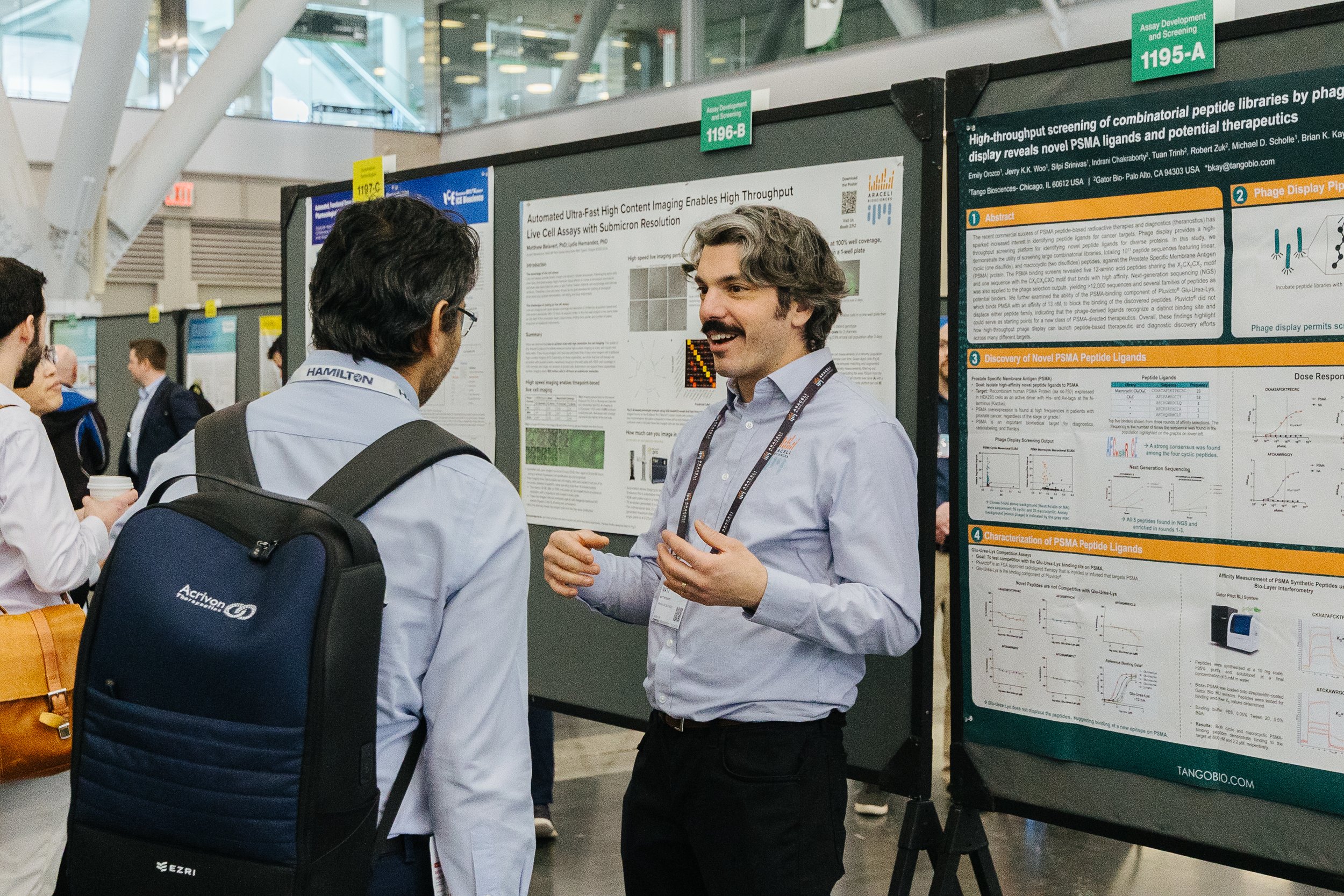 Two men are conversing at a scientific research poster presentation in a conference. The man on the right is explaining the poster to the man on the left. The poster is titled 'Automated Ultra-Fast High Content Imaging Enables High Throughput Live Cell Assays with Submicron Resolution' and contains scientific graphs and text. Other attendees are visible in the background.
