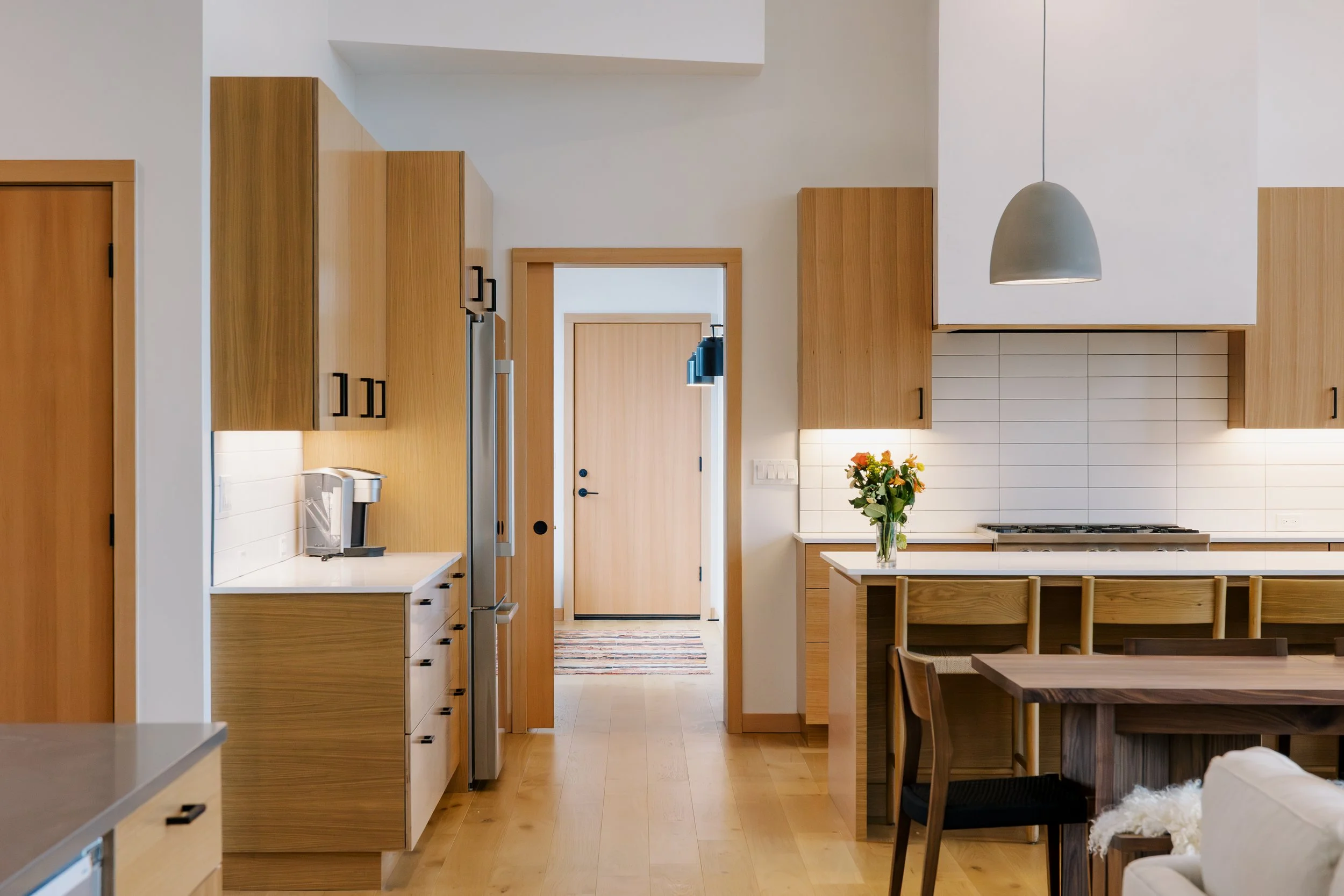 View of a modern kitchen with wooden cabinets, a white countertop, a vase of orange and yellow flowers, and an open doorway leading to another room.