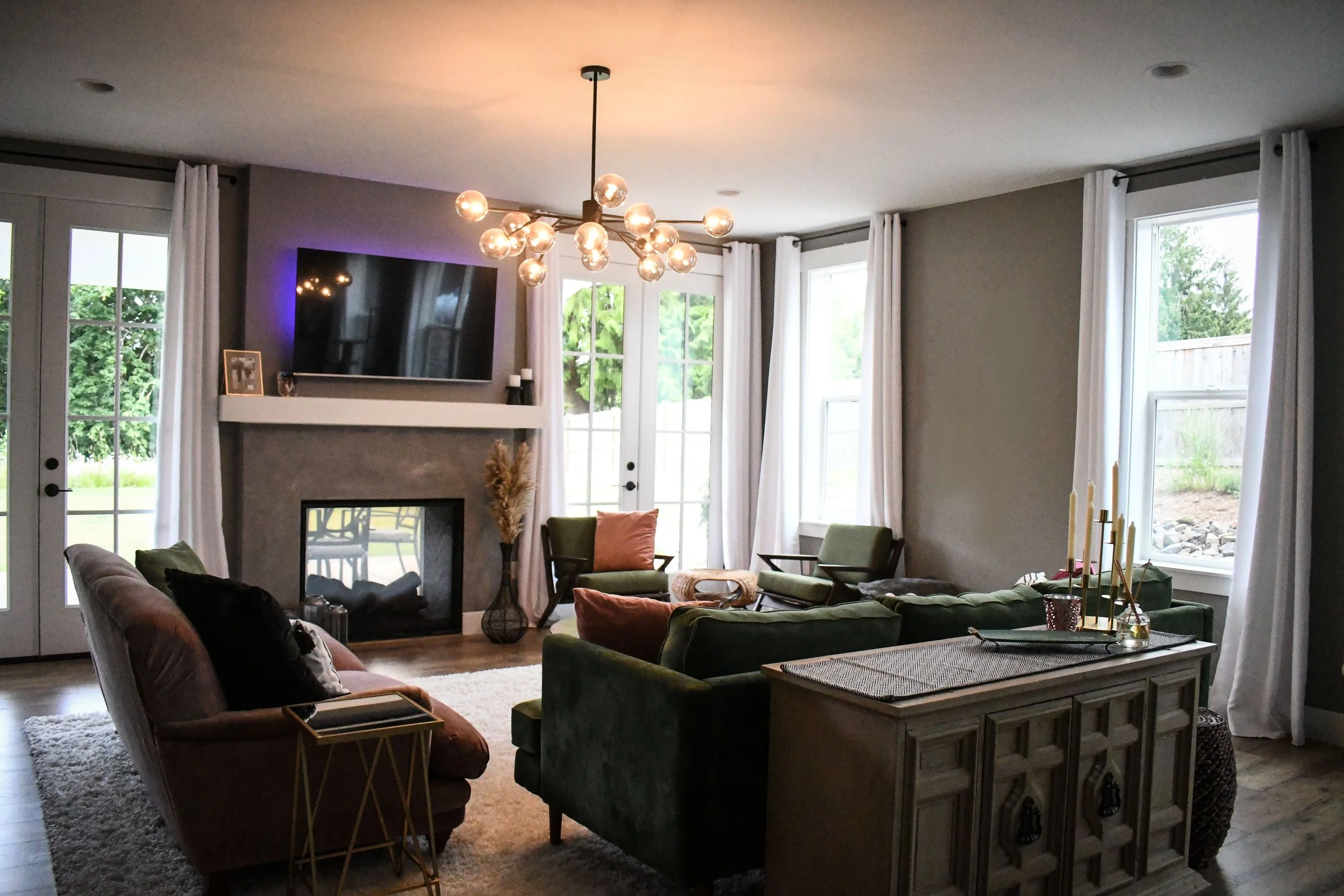Living room with green and pink sofas, a fireplace, a large TV above the fireplace, and large windows with white curtains, decorated with plants and candles.