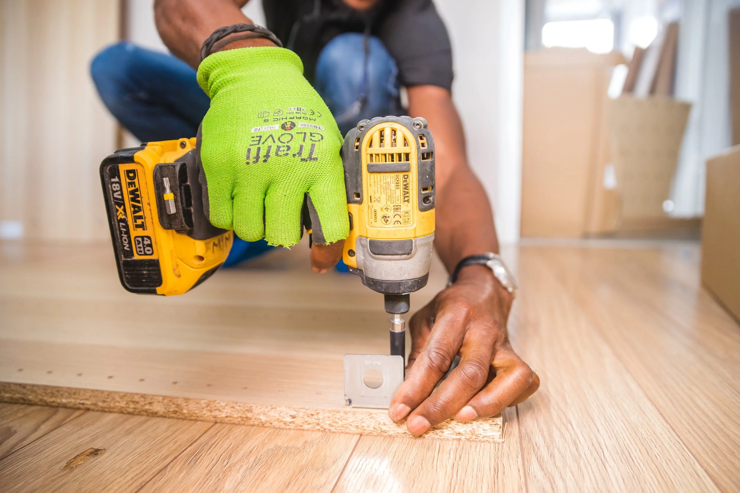 Person using a yellow DEWALT cordless drill with a green glove, fastening a metal bracket to a wooden surface during a woodworking project.