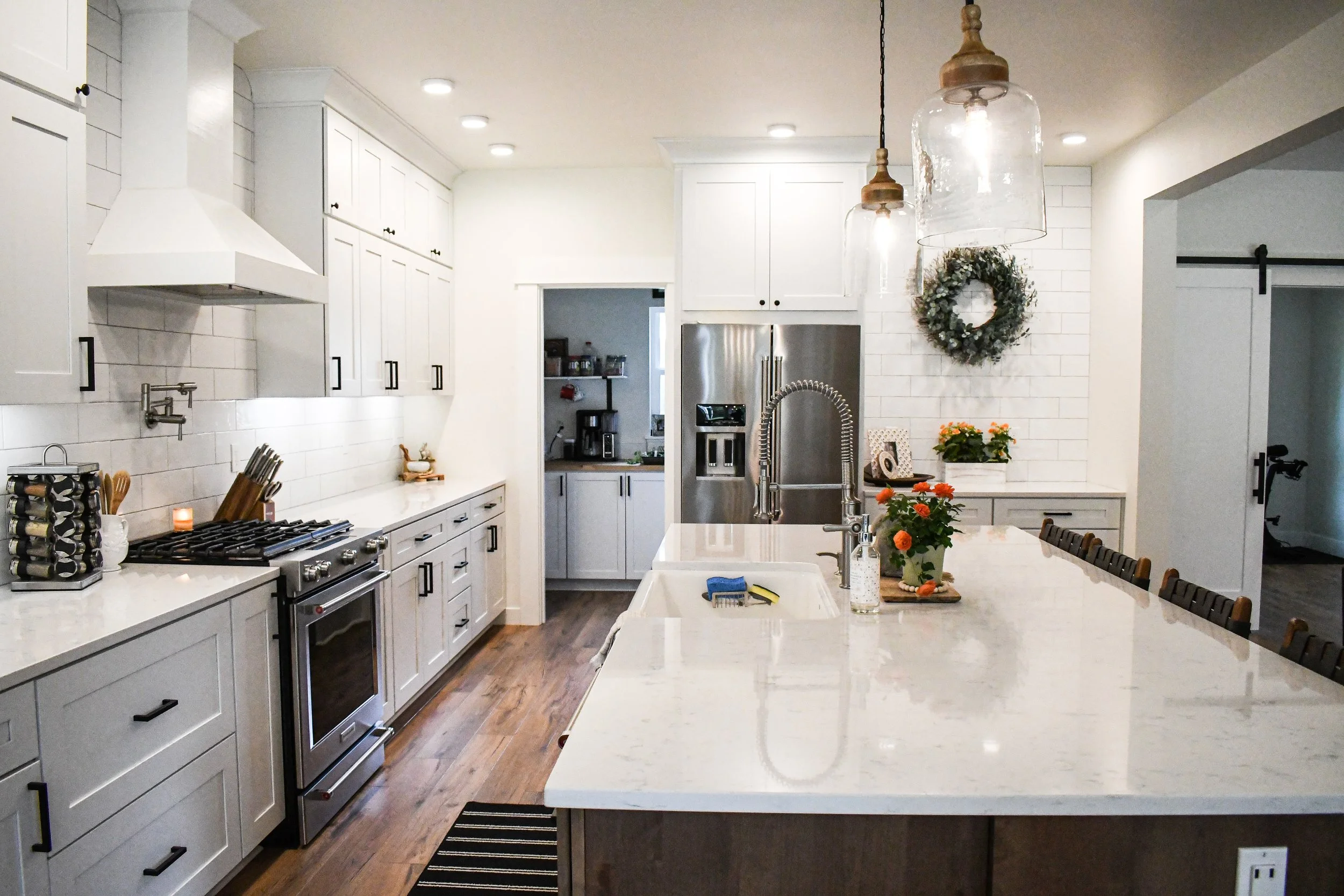 A modern kitchen with white cabinets and a large kitchen island with a marble countertop. There are pendant lights hanging above the island, a stainless steel refrigerator, and a stovetop with an oven. The kitchen is decorated with a wreath on the wall, potted flowers, and small decorative items. Wooden flooring is visible.