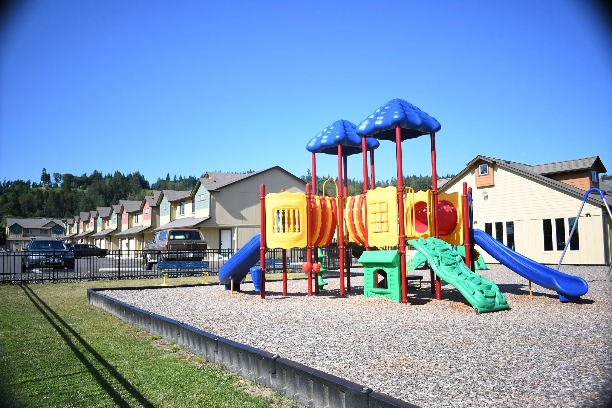 Colorful playground with slides and climbing structures in front of suburban neighborhood houses under a blue sky.