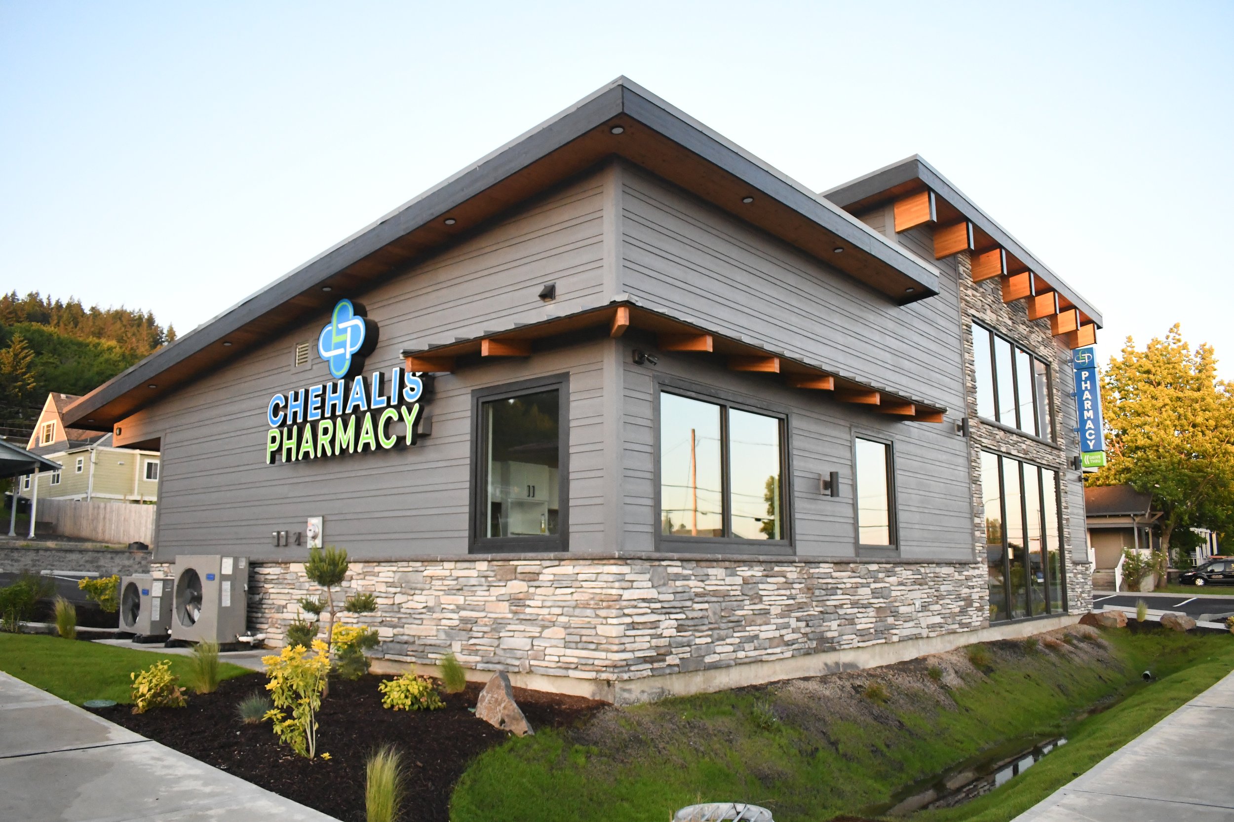 Exterior view of Chehalis Pharmacy with modern gray siding, stone accents, and large windows, surrounded by landscaped plants and lawns.