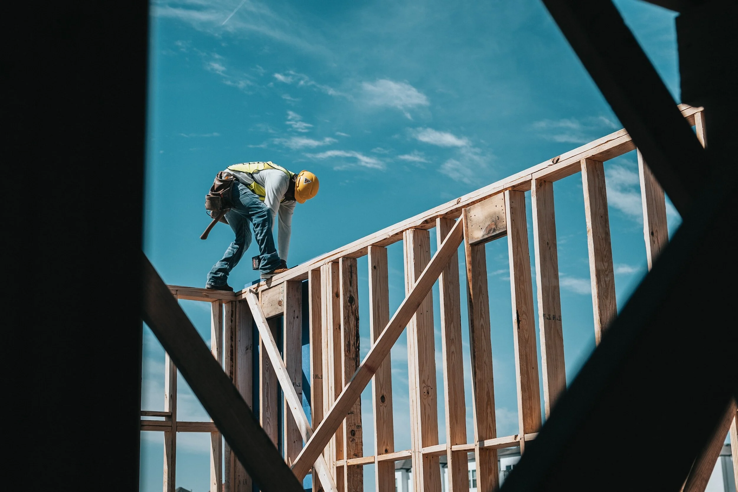 A construction worker wearing a yellow safety helmet and vest, working on the wooden frame of a building under a blue sky with scattered clouds.