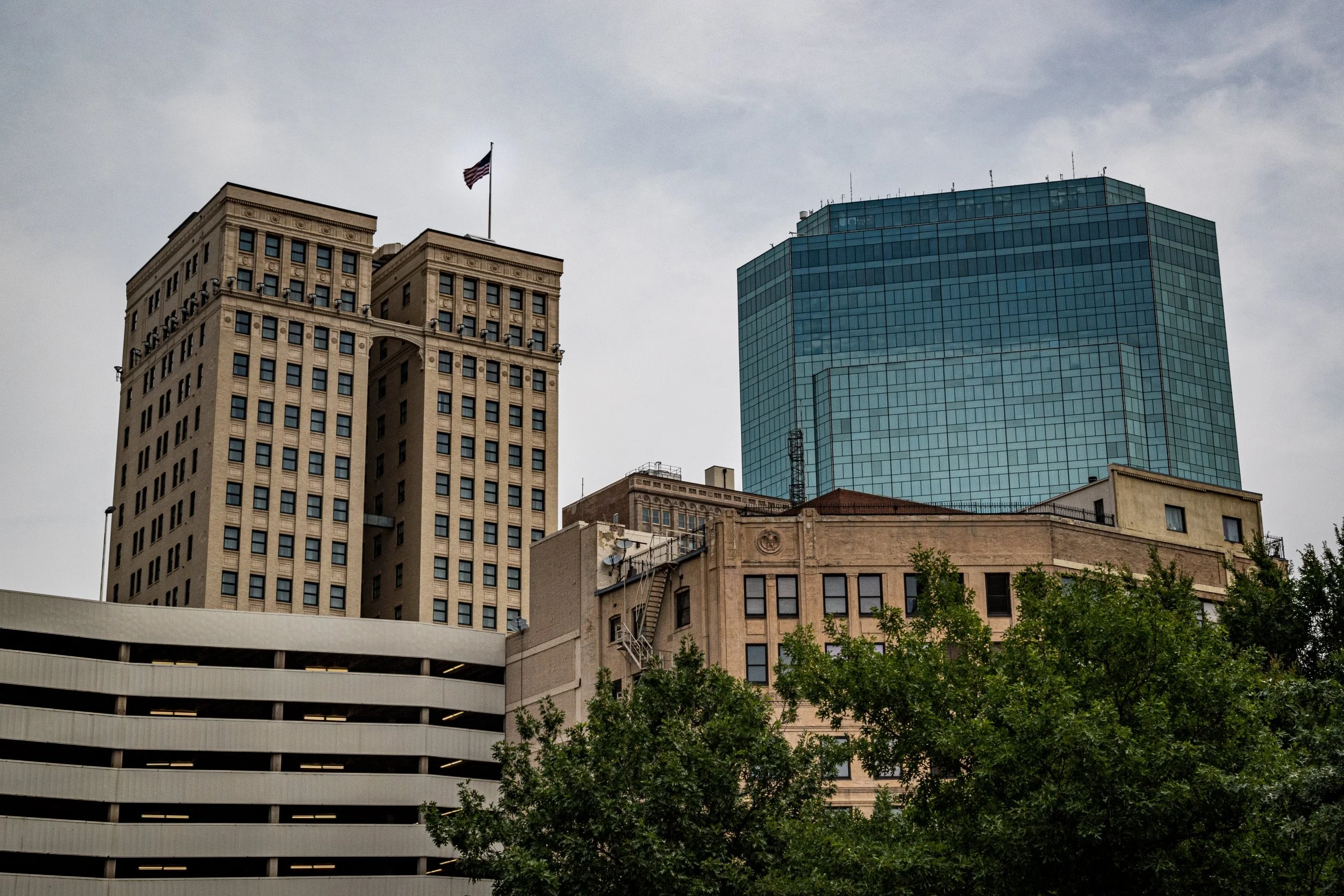 A city skyline featuring a beige skyscraper with an American flag on top, a modern blue glass building, and a parking garage with trees in the foreground.