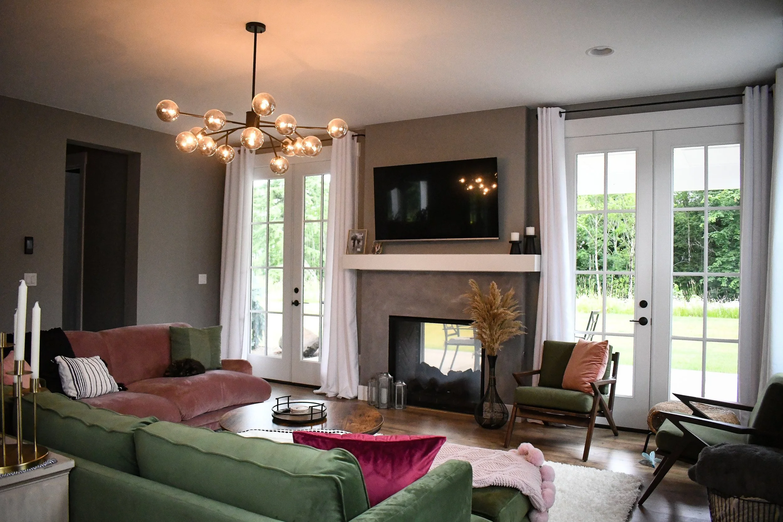 Living room with a pink velvet sofa, green armchair, and a fireplace with a TV above it. The room has large windows with white curtains, a wooden coffee table, and decorative items including candles, lanterns, and a vase with pampas grass. A modern c