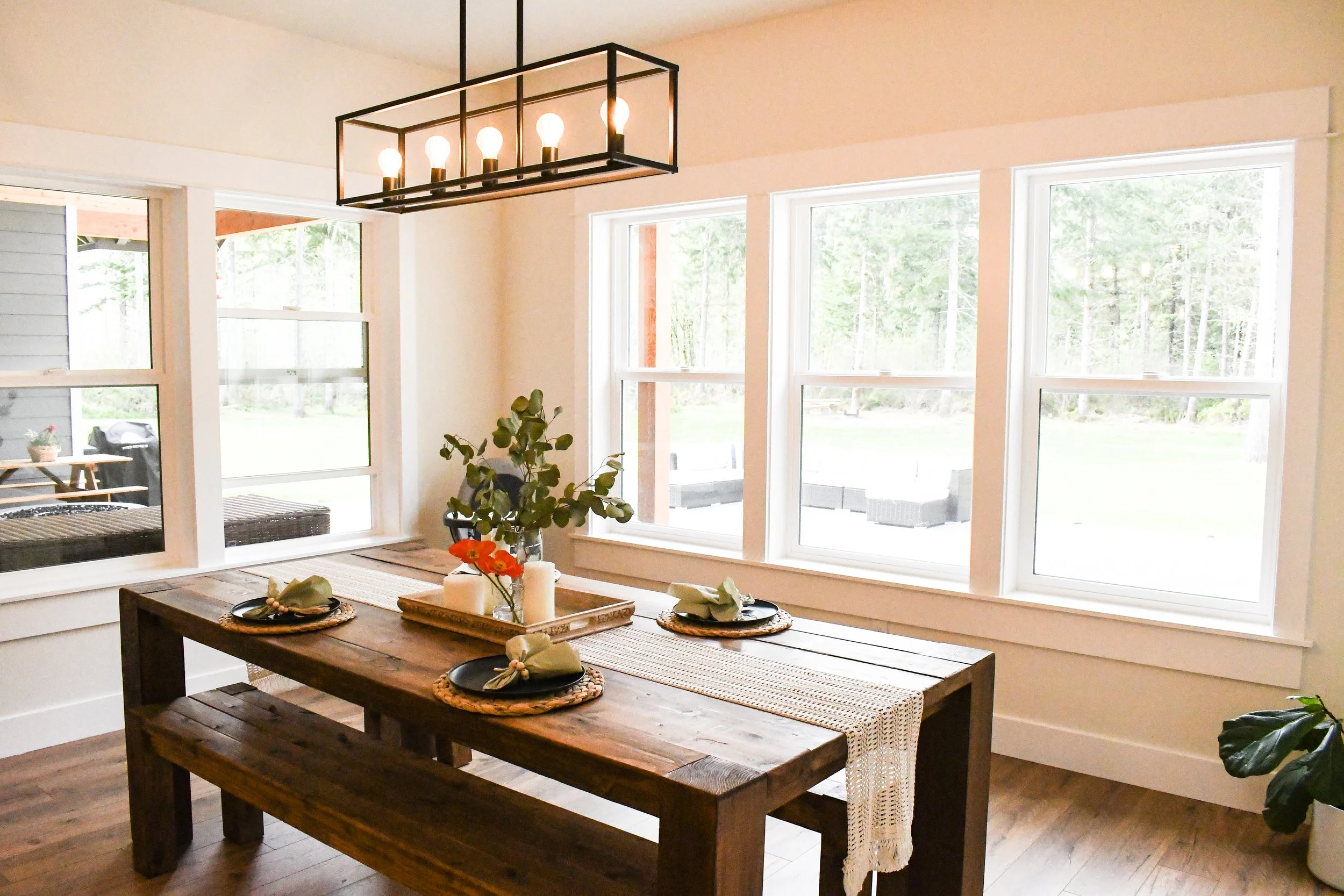 Dining room with a wooden table set with black plates, napkins, and silverware, decorated with candles and greenery. Large windows provide natural light and a view of the backyard, with a modern light fixture hanging above the table.