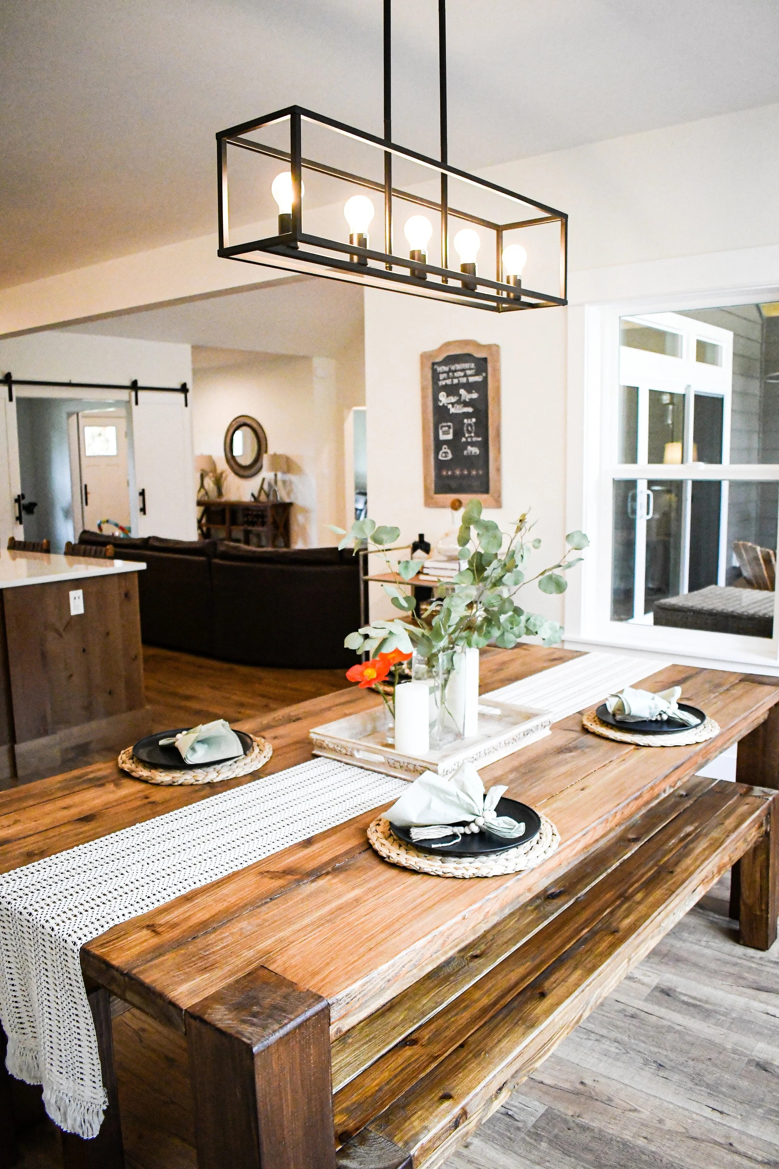 A dining table set for three, with placemats, napkins, and black plates, in a home with a rustic, wooden interior and a modern black chandelier.