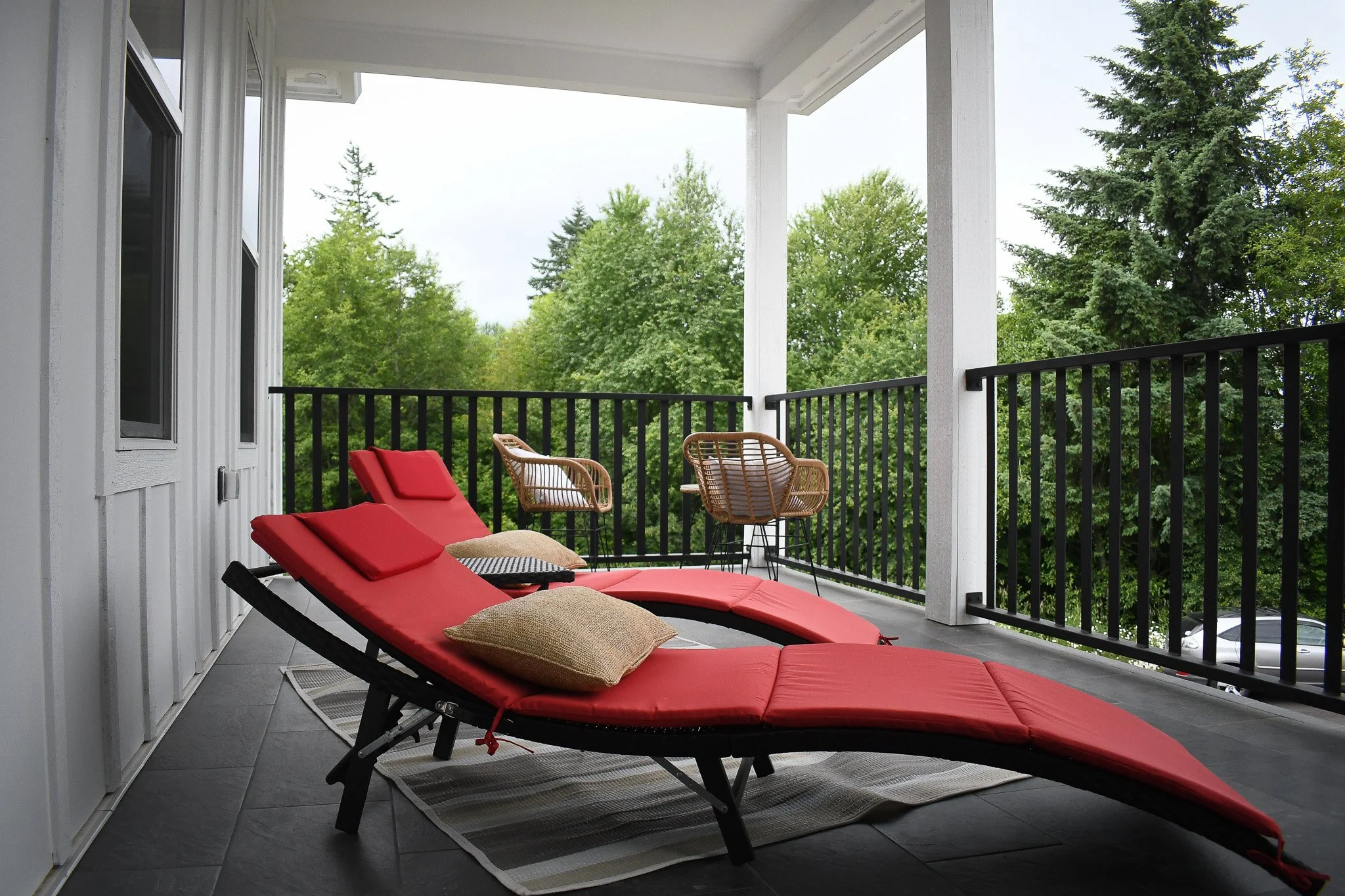 Balcony with two red lounge chairs, pillows, and wicker chairs overlooking green trees.