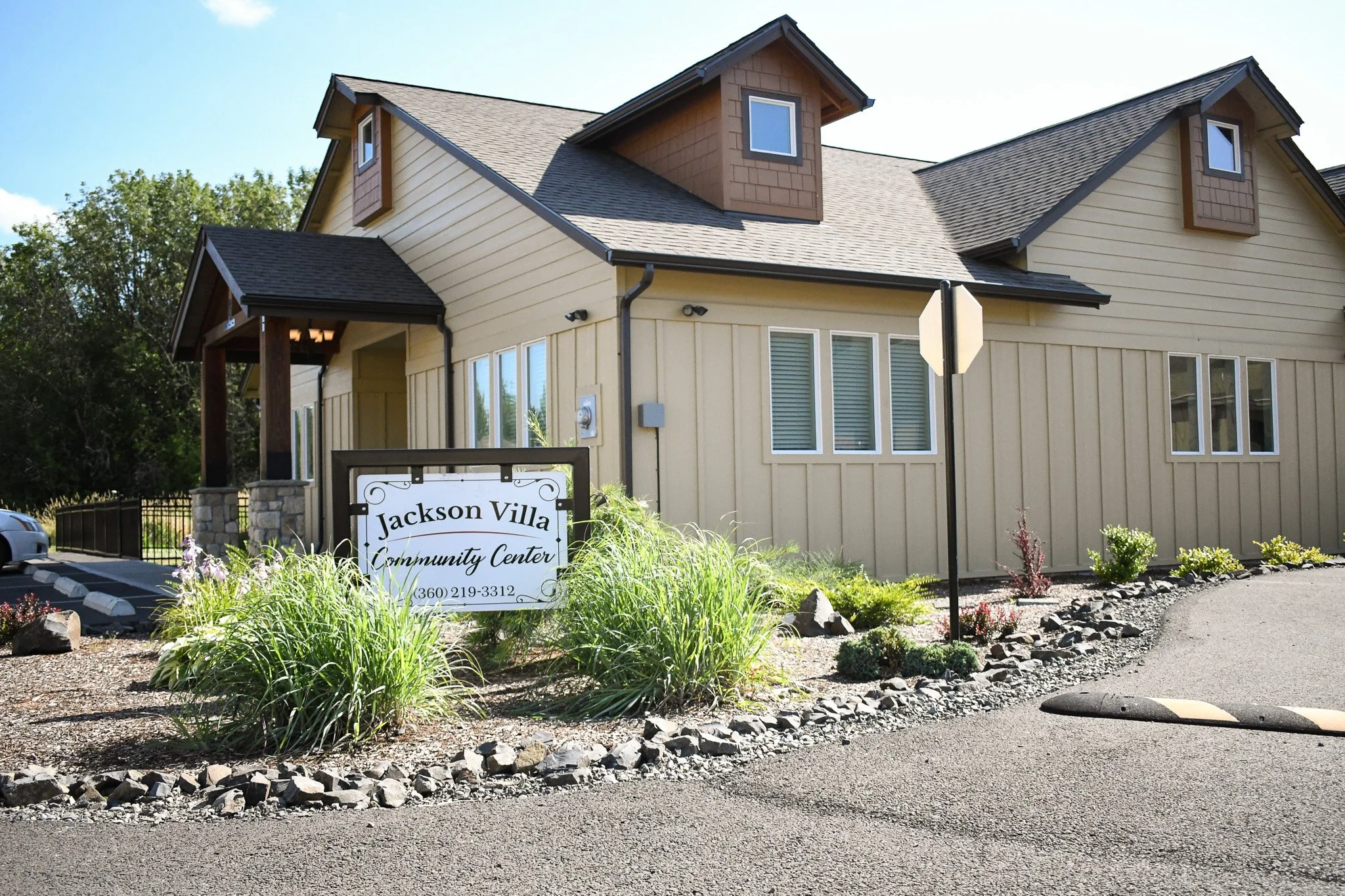 Exterior of a beige multi-story community center building with a sign that reads "Jackson Villa Community Center" in front, surrounded by a landscaped garden with bushes and rocks, and a parking lot with a parked car.