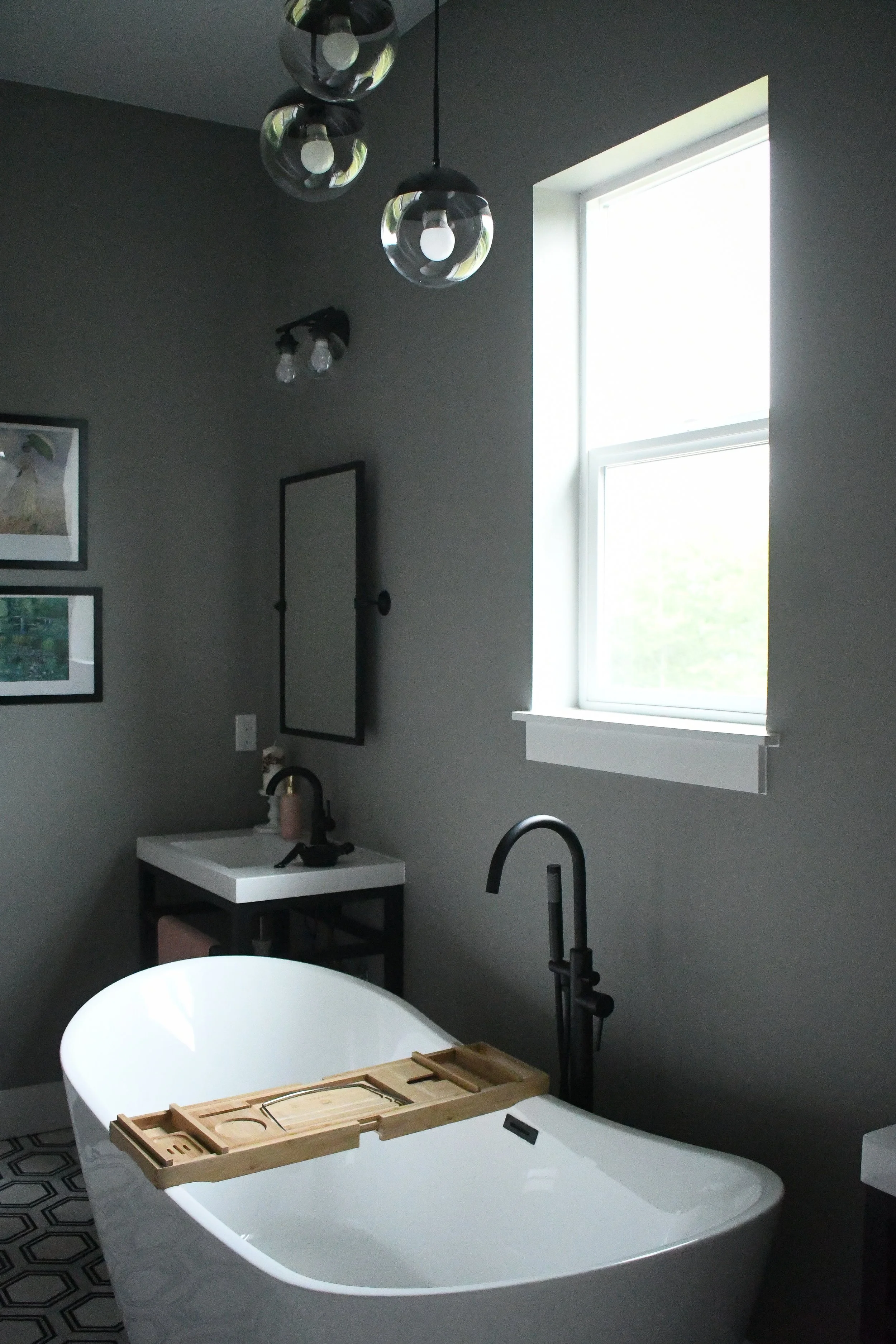 A modern bathroom with a freestanding bathtub, black fixtures, a window allowing natural light, and a gray wall with framed pictures.