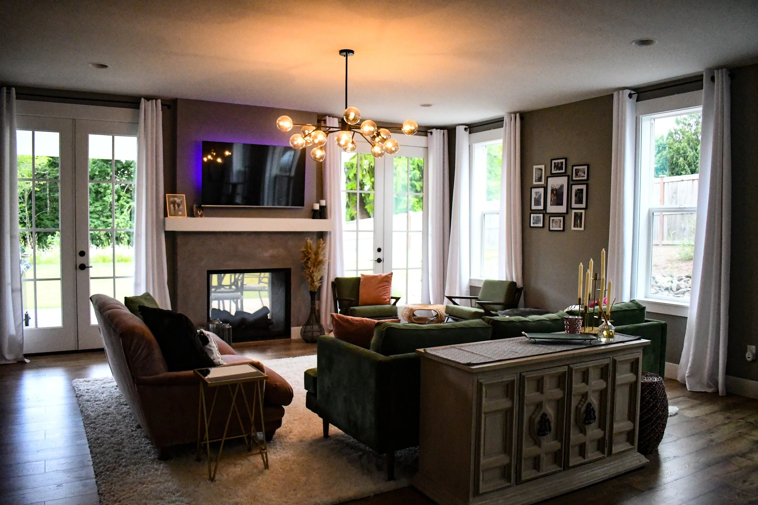 Living room with large windows, a fireplace, a wall-mounted TV, a chandelier, green and orange chairs, a gray couch, a patterned sideboard, and wall art