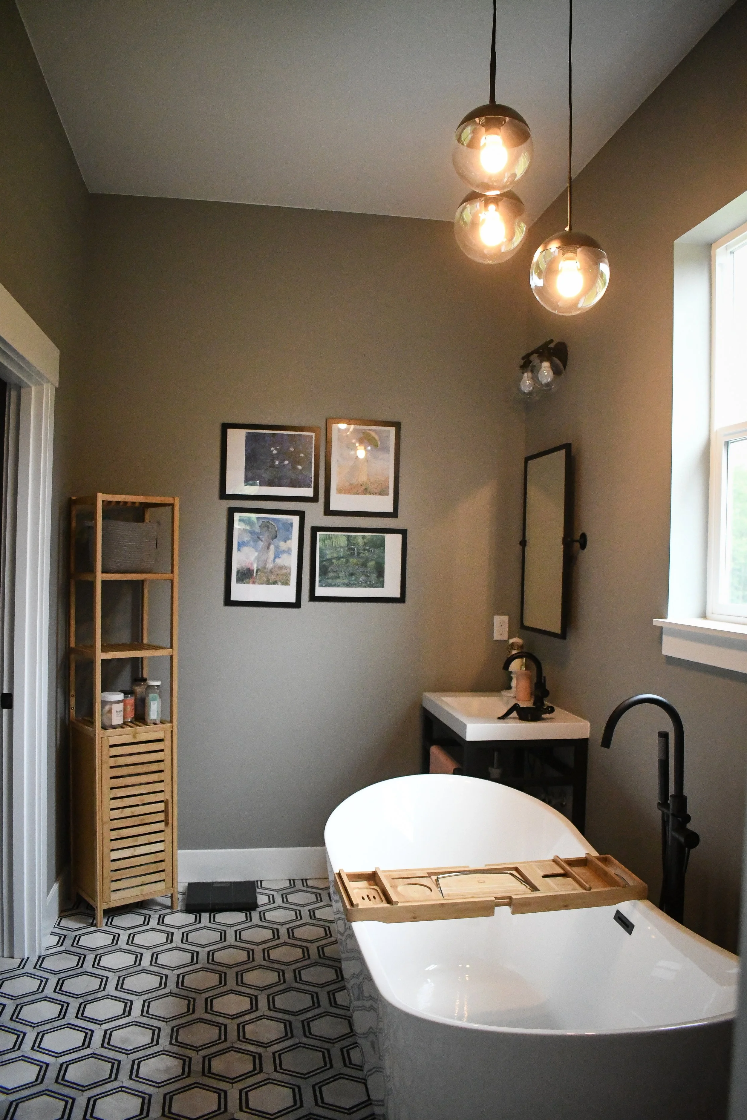 A bathroom with a freestanding bathtub, a small sink with a mirror, a wooden shelf with toiletries, and a set of four framed pictures on a gray wall. There are modern pendant lights hanging from the ceiling and a window providing natural light.