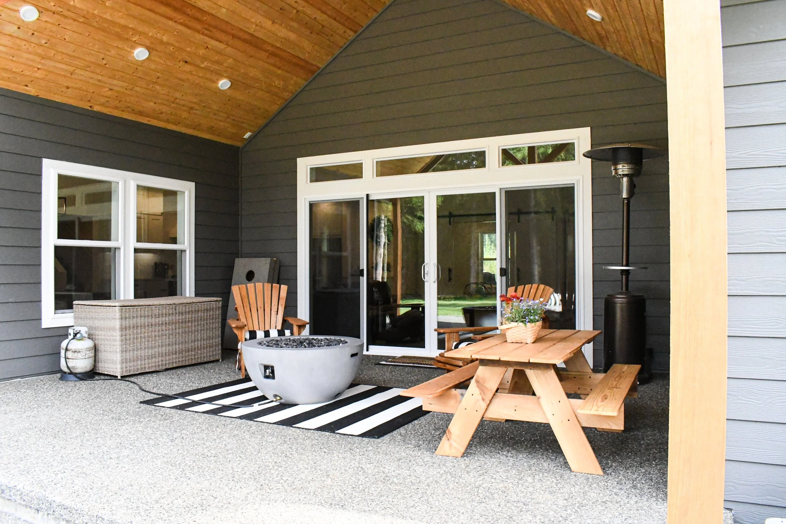 Covered outdoor patio with wooden ceiling, sliding glass door, wicker storage box, wooden chair, fire pit on a black-and-white striped rug, wooden picnic table with flower basket, patio heater, and dark gray siding house with white window frames.