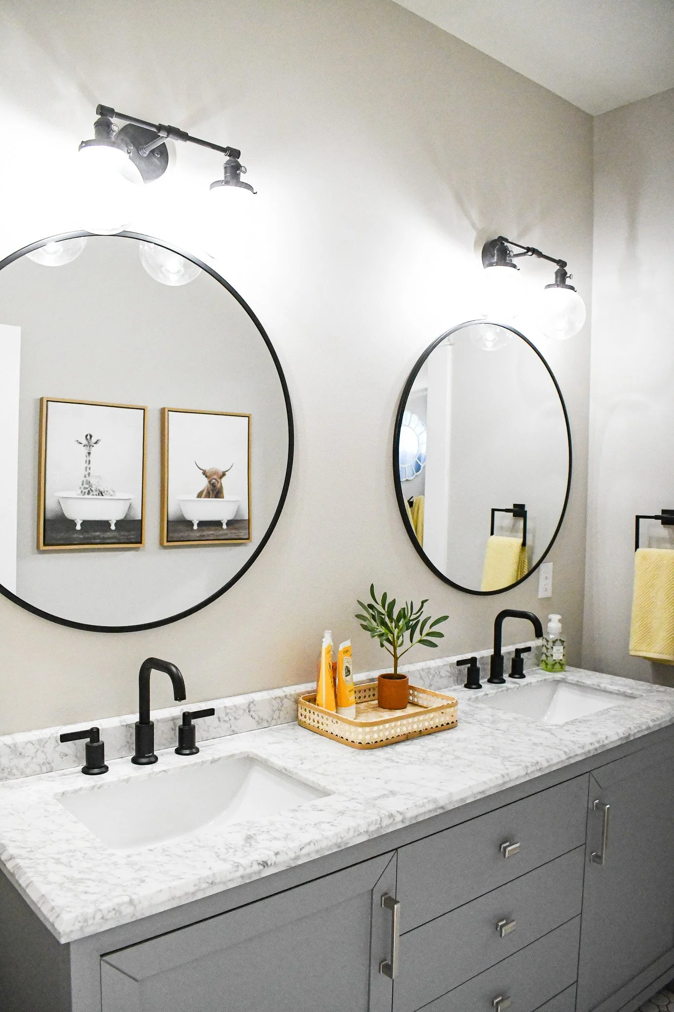 Bathroom with double vanity, marble countertop, oval mirrors, black fixtures, framed animal artwork, and yellow towels.