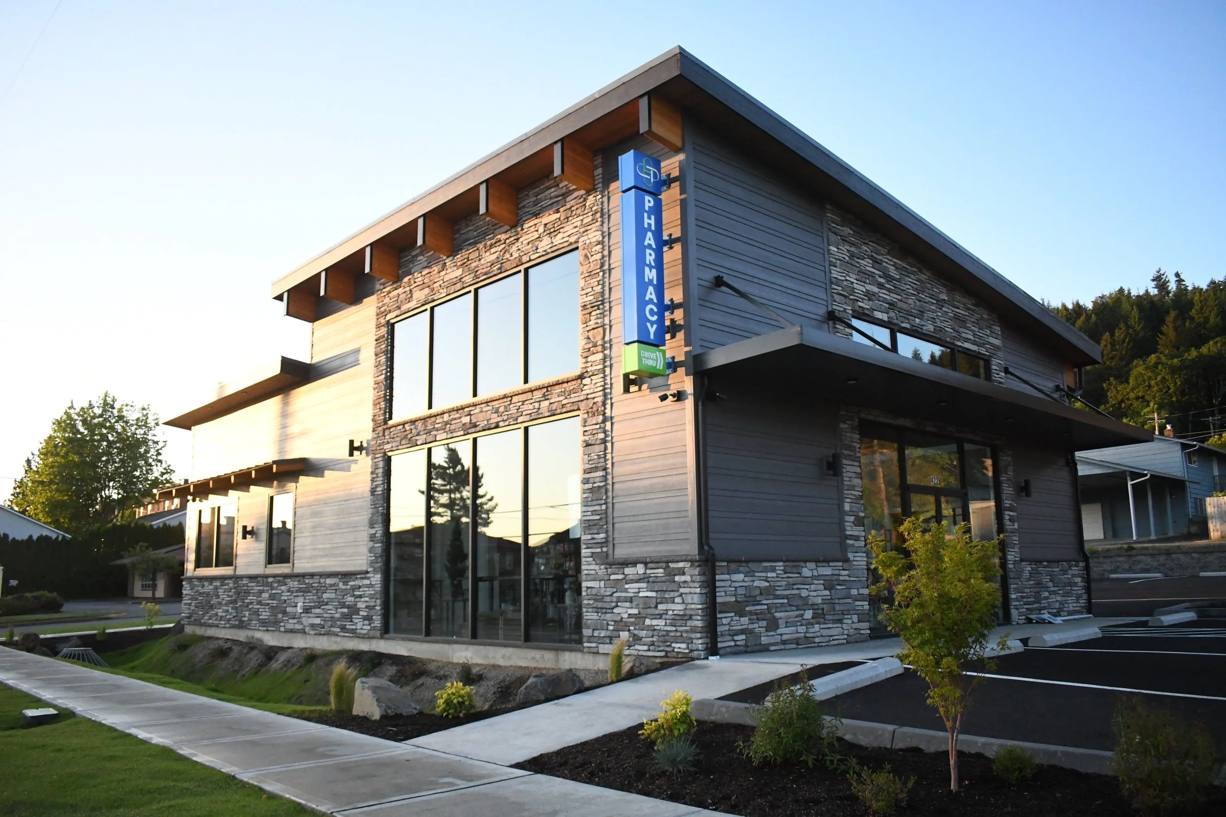 Modern two-story pharmacy building with large glass windows, stone and wood siding, and a blue vertical pharmacy sign