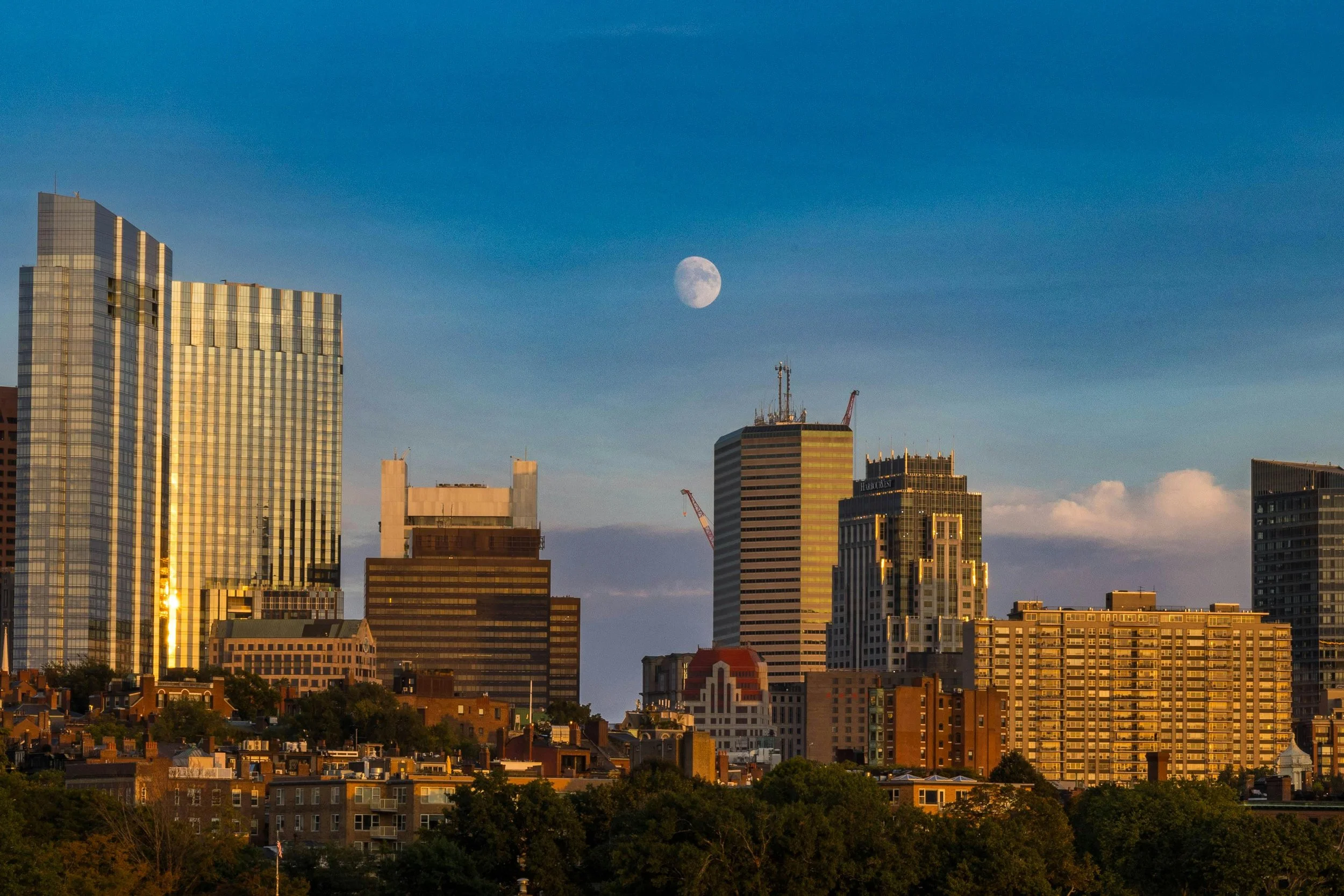 City skyline with tall buildings during sunset, with the moon visible in the evening sky.