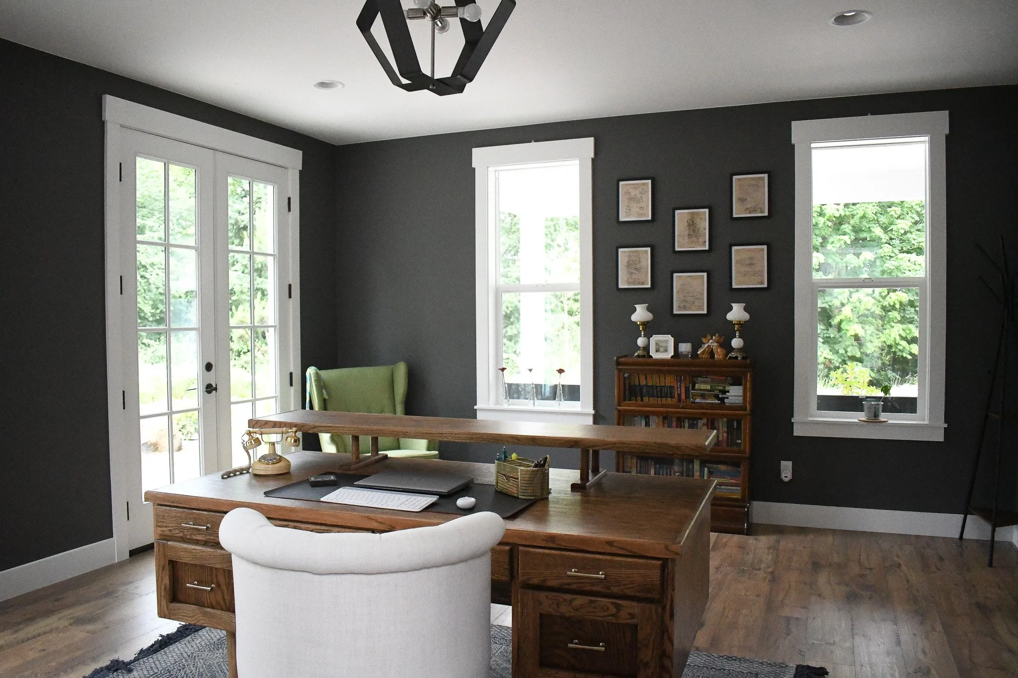 A home office with a dark gray wall, white trim, and three windows. There is a wooden desk with a white chair and a green armchair in the corner. A bookshelf with lamps and decorative items is against the wall. The room is well-lit with natural light.