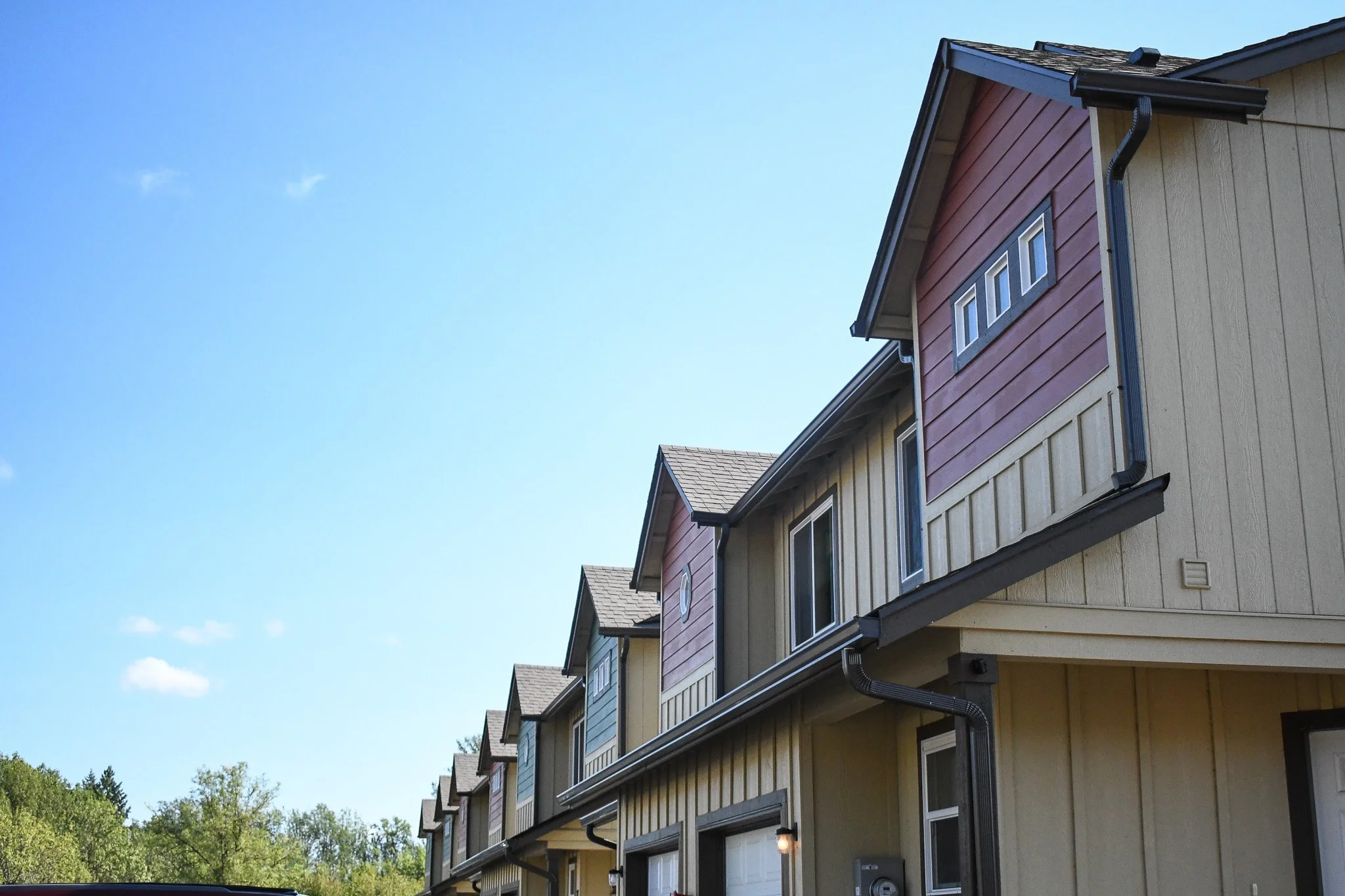 Row of modern townhouses with colorful siding under a blue sky, trees in the background.