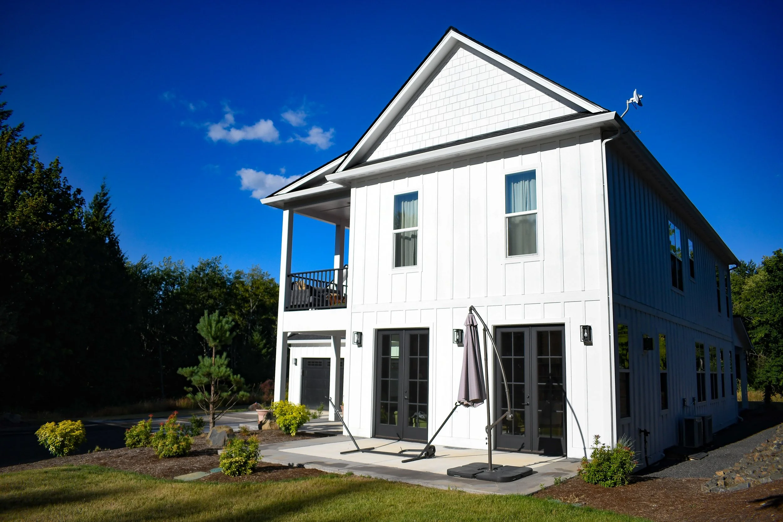 A modern white two-story house with black-trimmed glass doors and windows, a small balcony, outdoor workout equipment, and landscaped yard with bushes and trees under a bright blue sky.