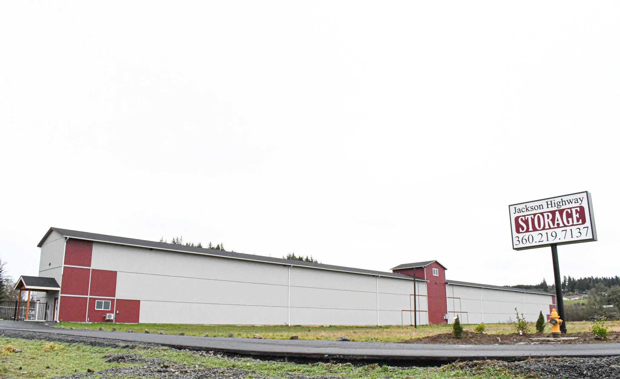 Large rectangular storage building with red accents and a sign that says 'Jackson Highway Storage' with a phone number, grass and a road in the foreground.