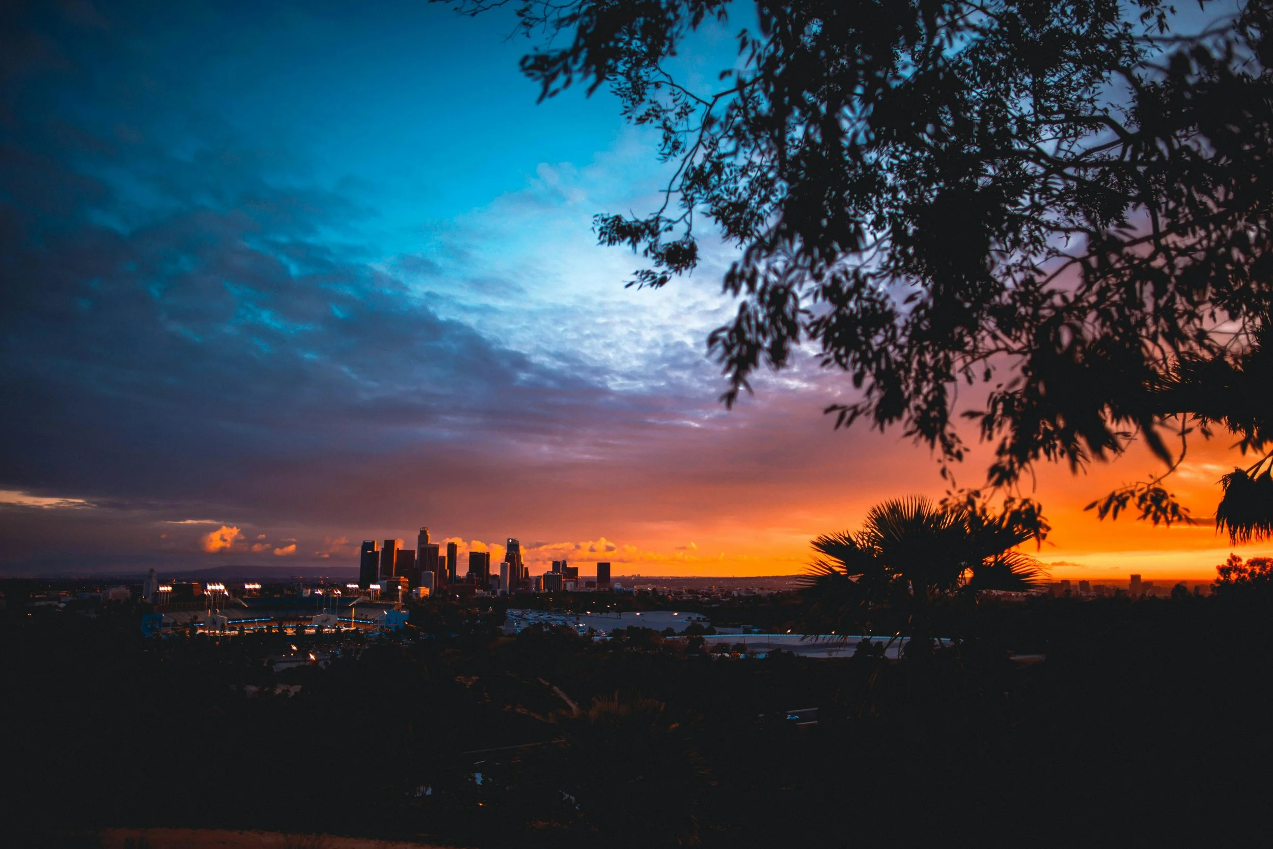 Sunset over a city skyline with silhouettes of trees and clouds in the sky.