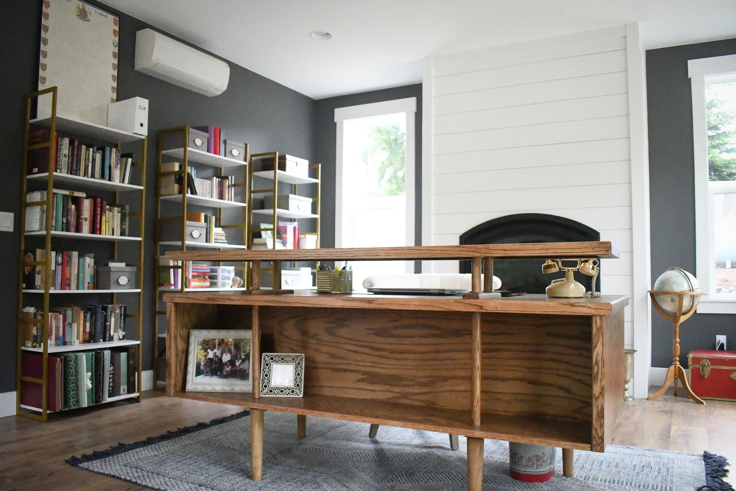 A home office or study room with a large wooden desk, bookshelves filled with books, and a fireplace with a white shiplap wall. There are two windows letting in natural light, and various decorative items such as picture frames, a globe, an old rotar