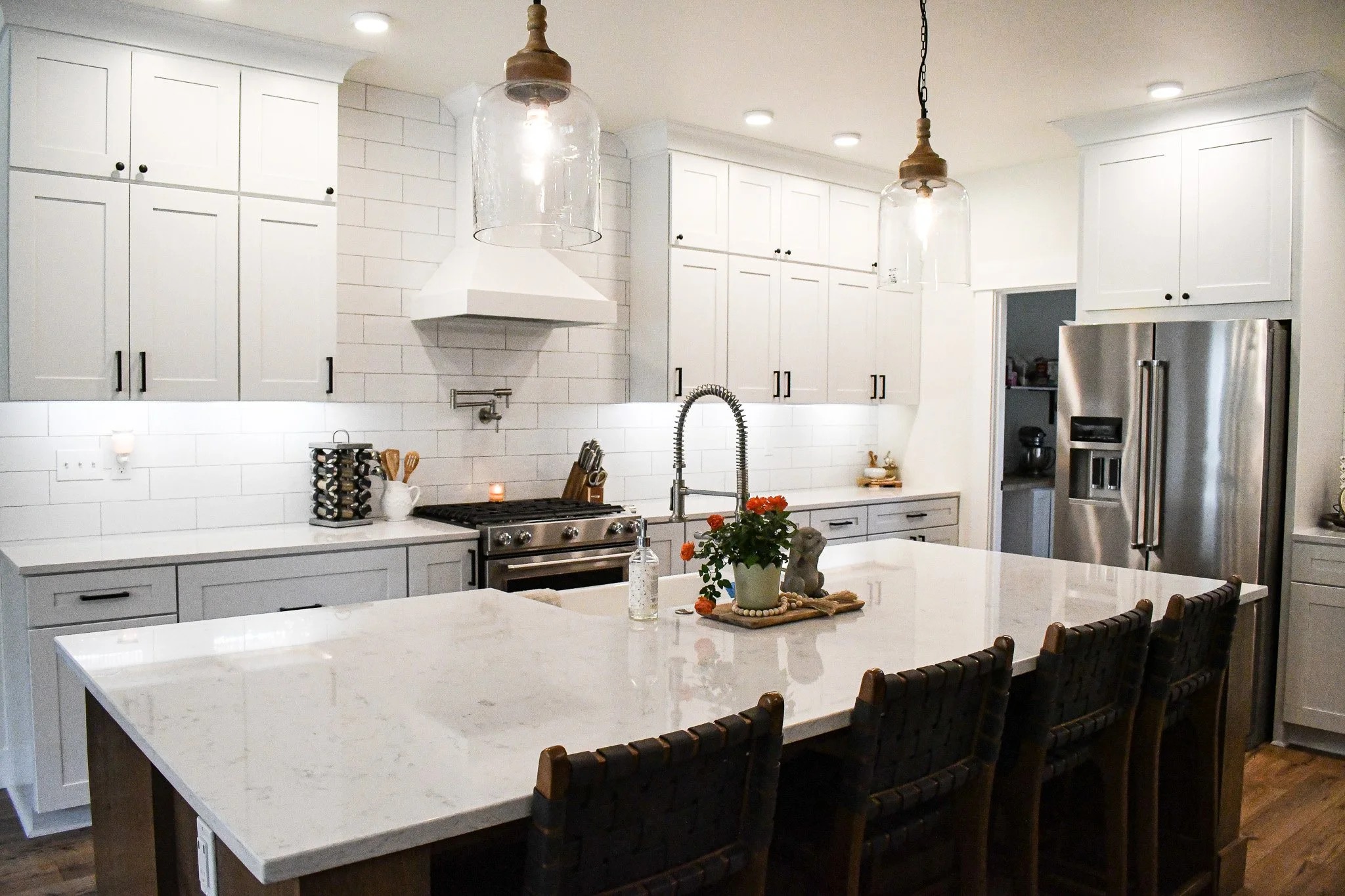 Modern kitchen with white cabinets, stainless steel appliances, and a large marble island with dark woven chairs. Pendant lights hang above the island, and decorative items like a potted plant and soap dispenser are on the island. The kitchen feature