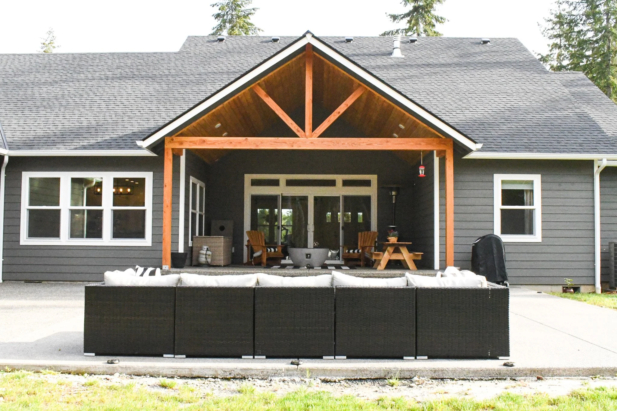 Back patio of a house with outdoor seating, a fire pit, and a covered wooden porch area with chairs, a picnic table, and a barbecue grill.