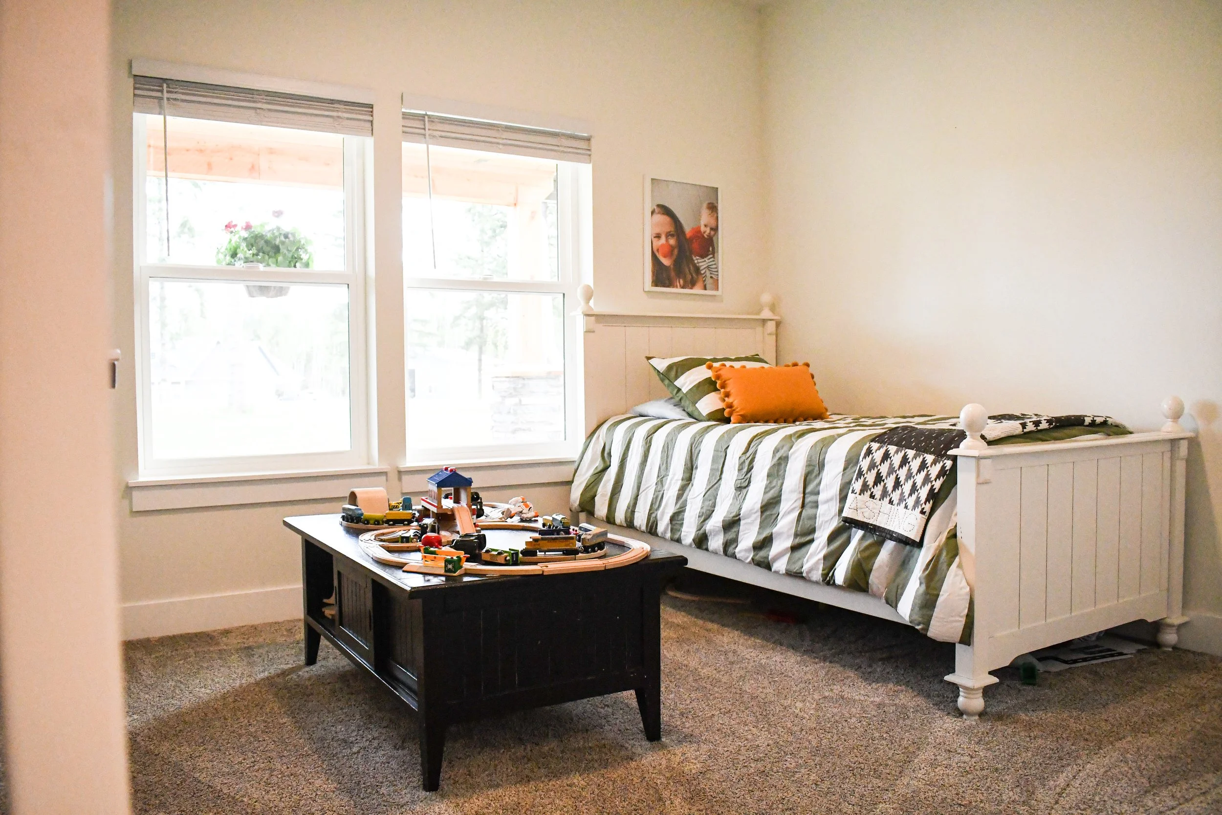 Children's bedroom with white bed, striped green and white bedding, orange pillows, toy train table, large window, and wall decoration of a woman and child.