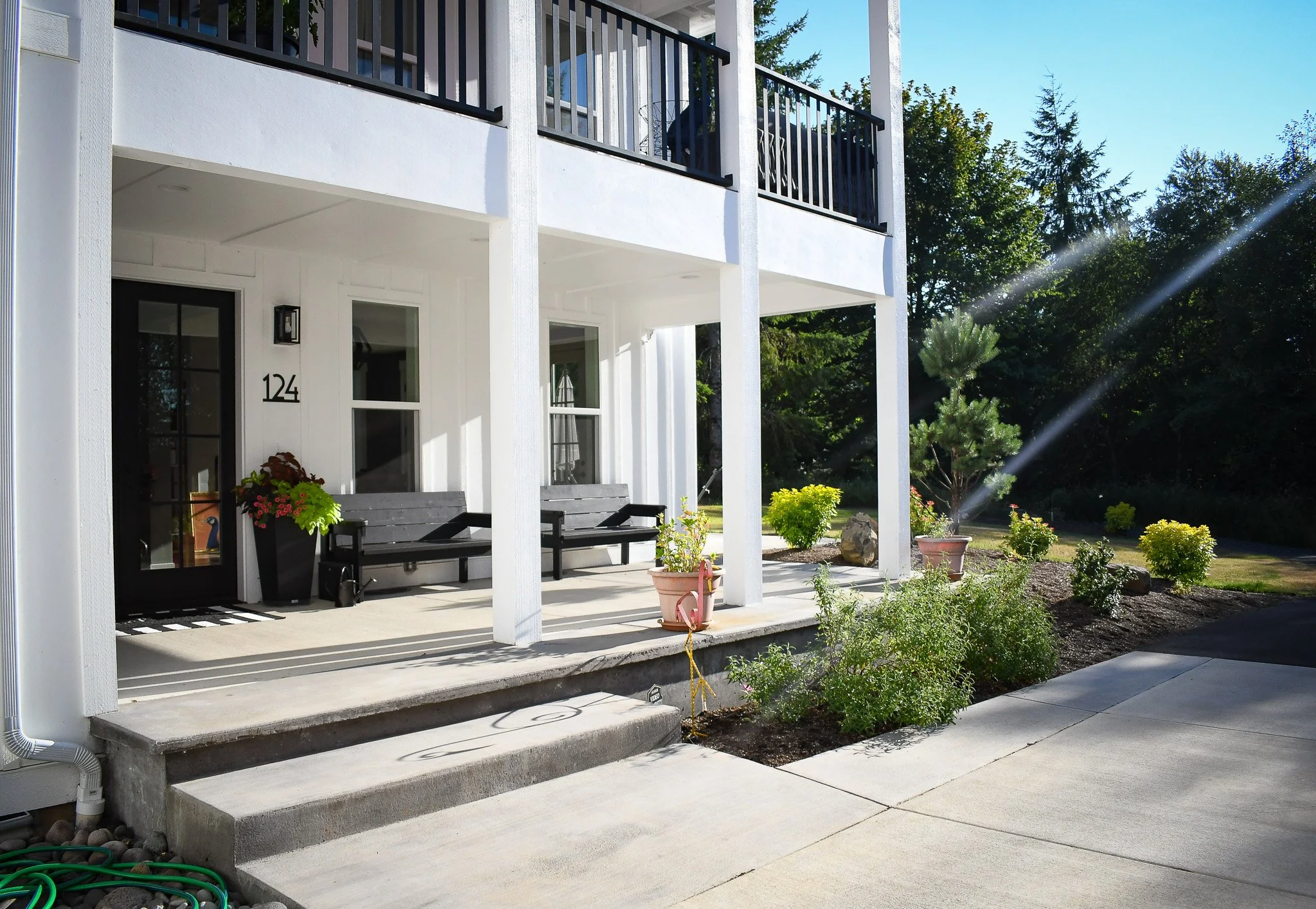 Front porch of a modern white two-story house with black accents, outdoor seating, potted plants, and a garden with small trees and bushes.