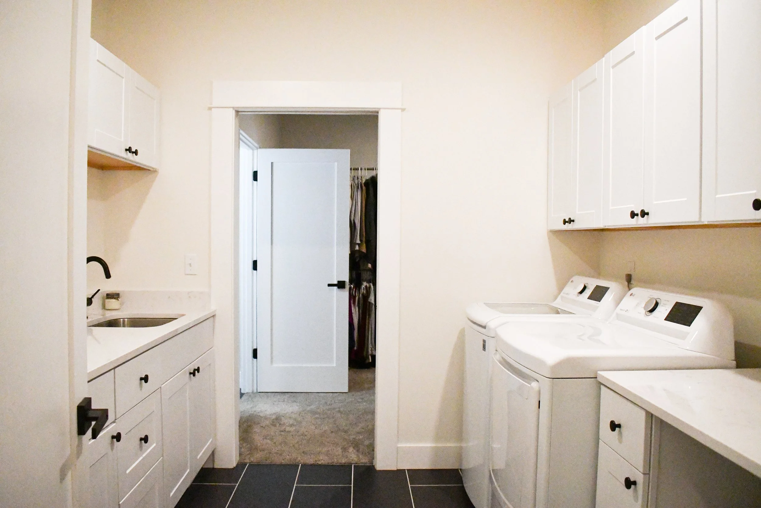 A laundry room with white cabinets, a washer and dryer, a small sink, and a doorway leading to a walk-in closet with clothes.